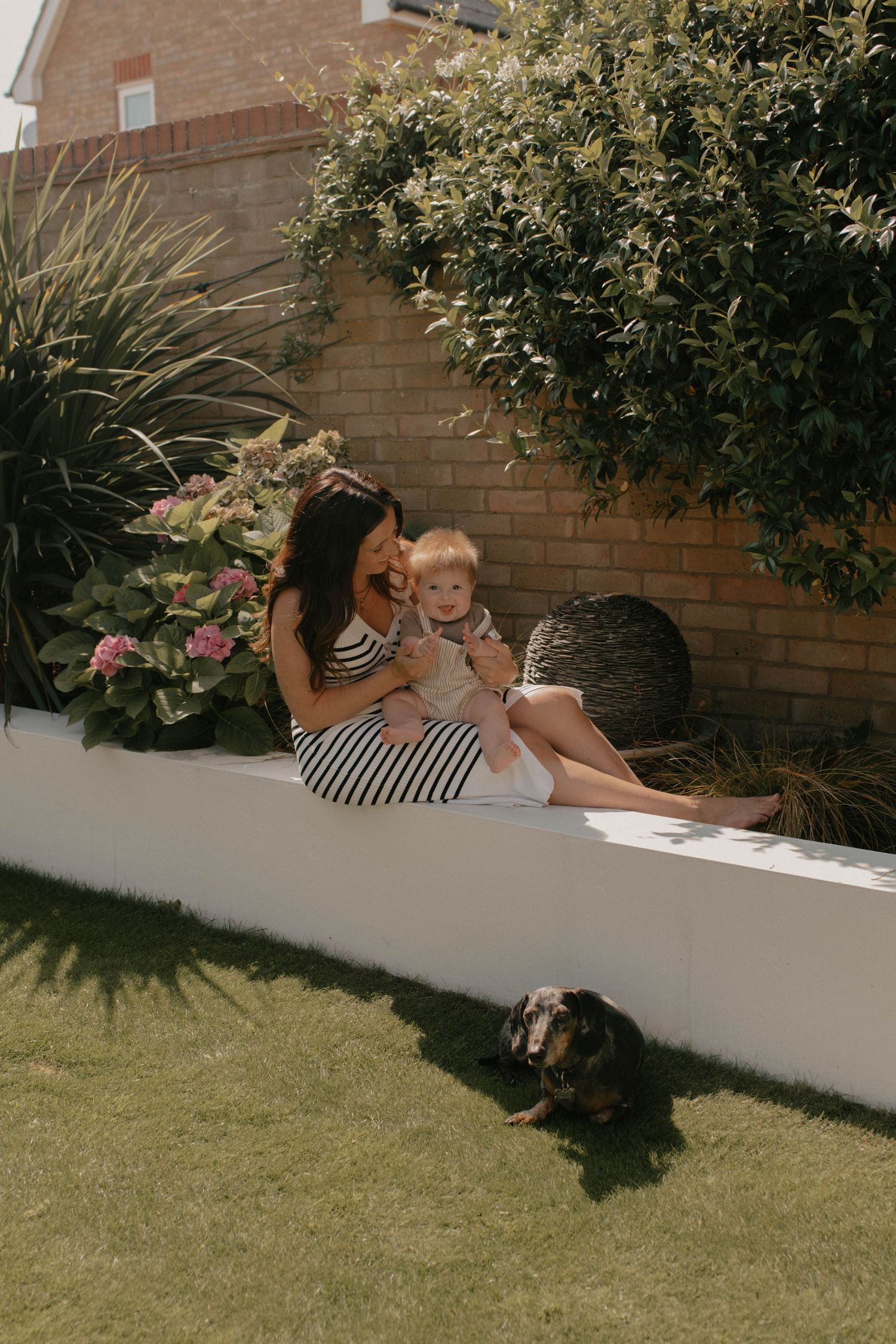 People relaxing on a white garden wall with lush greenery and brick backdrop on a sunny day.