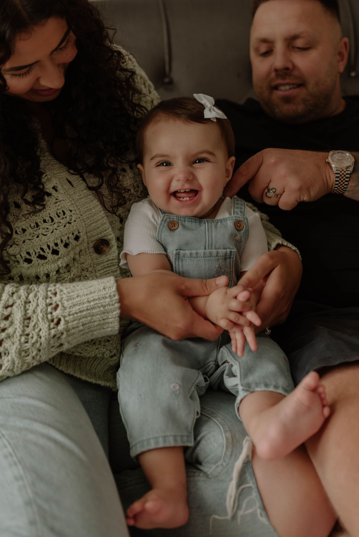 Joyful baby wearing denim overalls and hair bow laughs while being held by family members in cozy indoor setting.