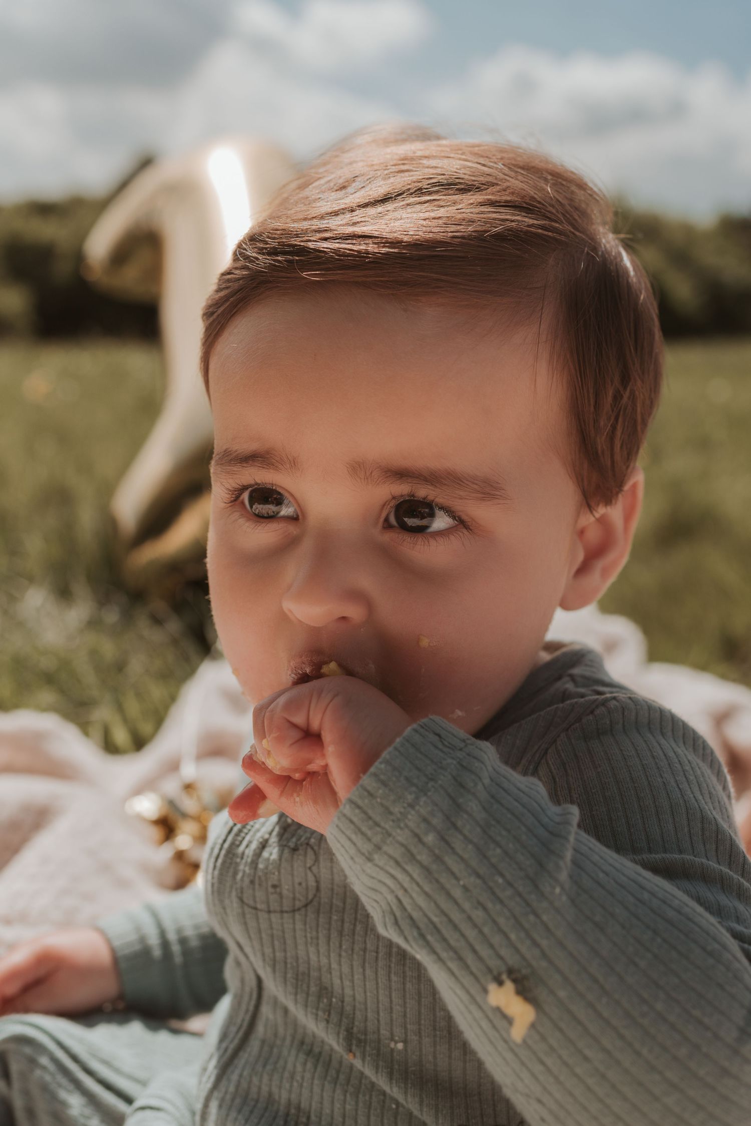 Child wearing a gray sweater nibbles on birthday cake while relaxing outdoors.