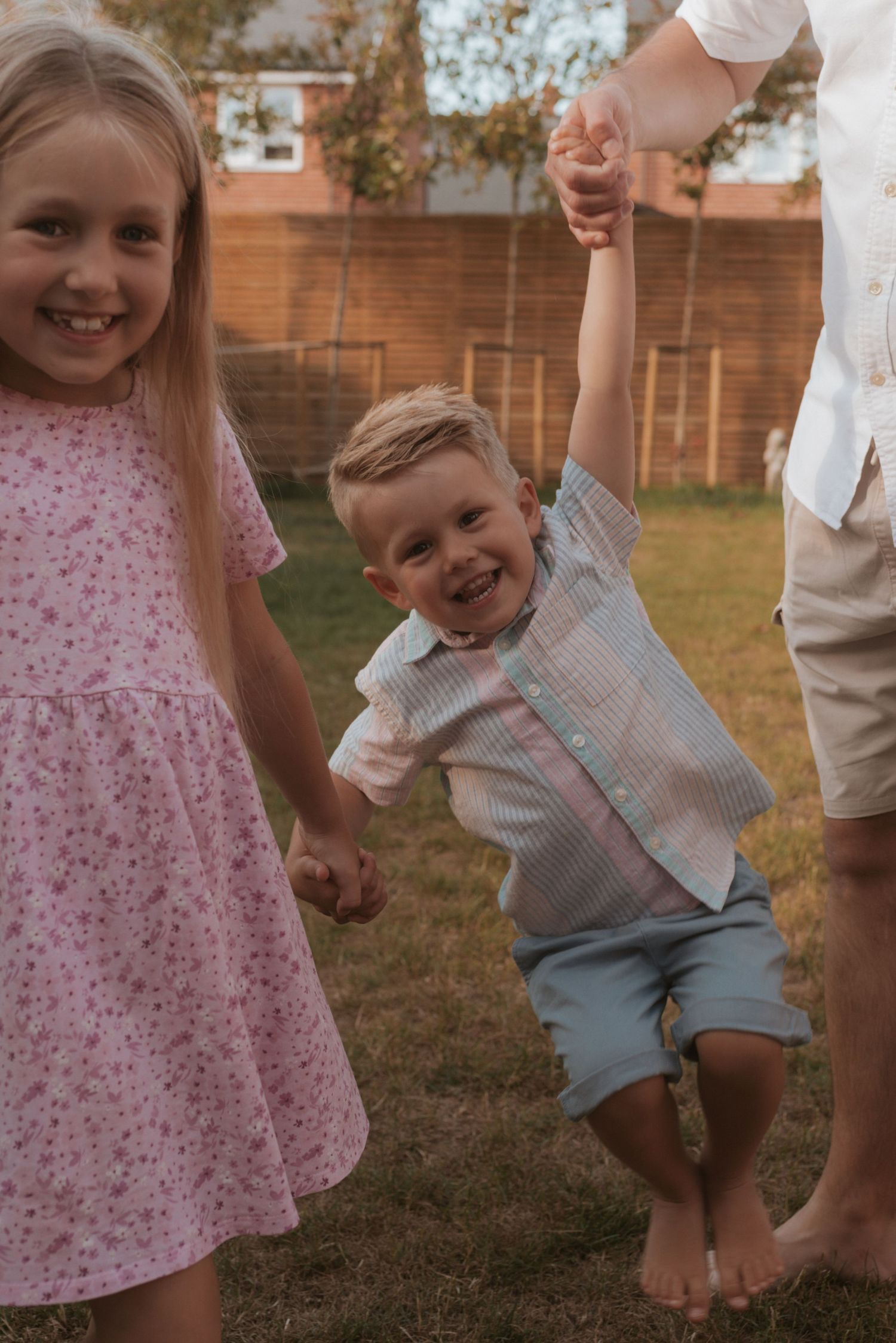 Children play together outdoors on a summer evening, with one jumping joyfully while holding hands in a backyard.