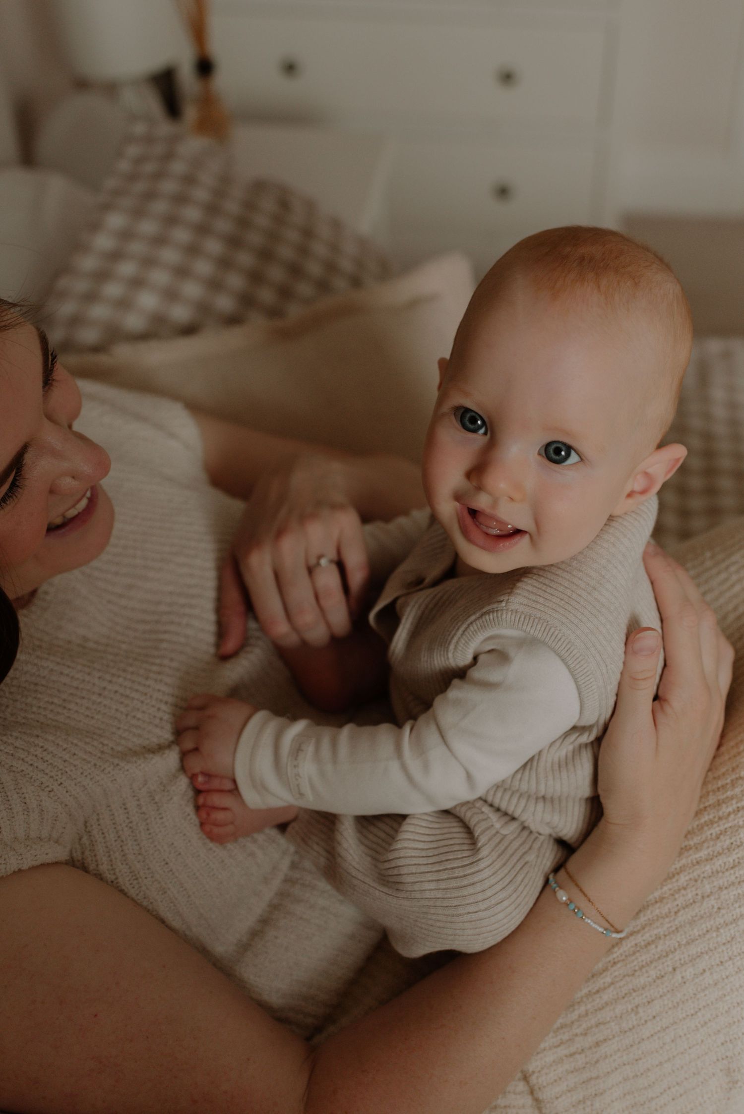 A baby in a white knit outfit lies on a textured beige blanket while being gently touched by caring hands.
