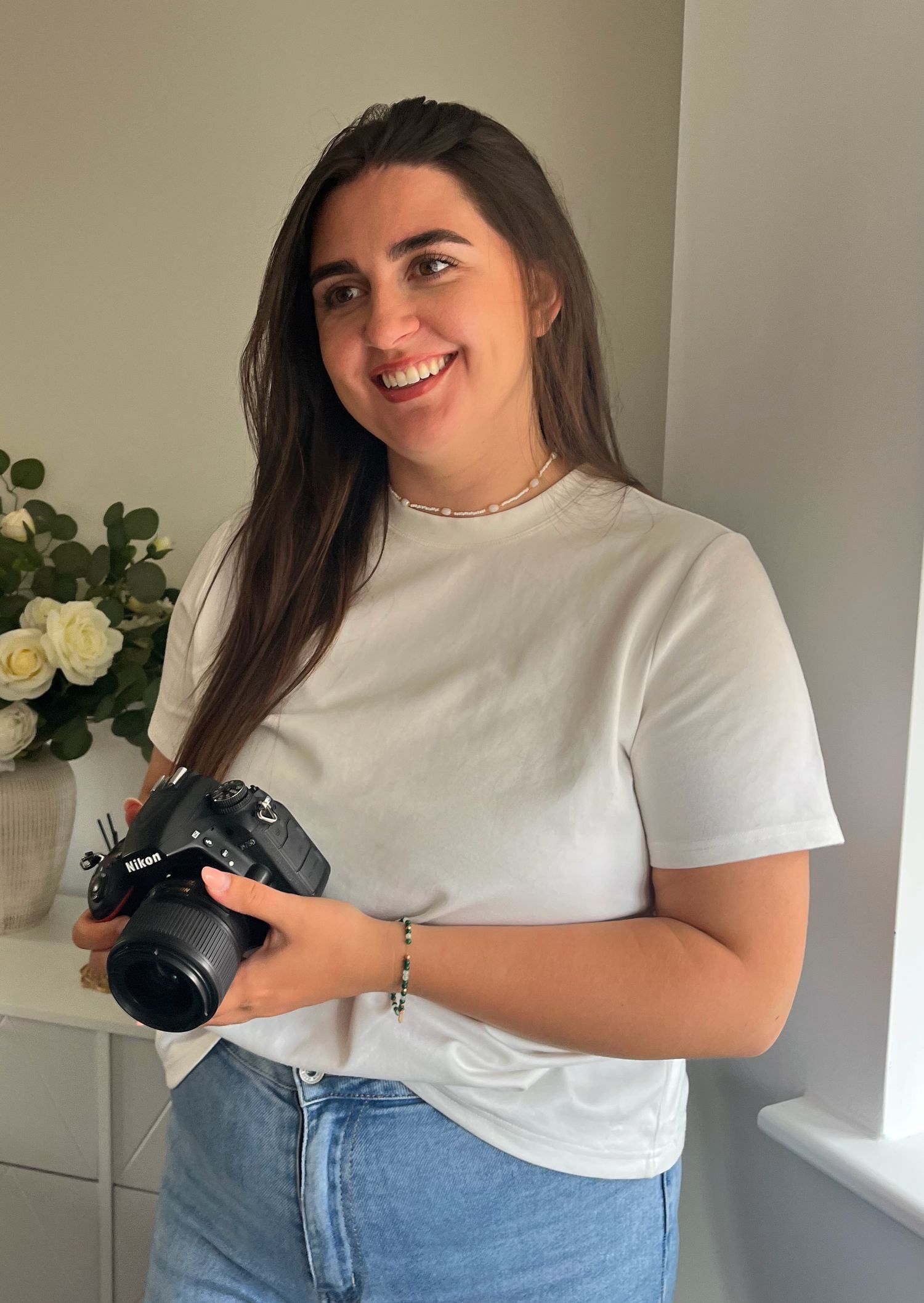 A person in a white t-shirt and jeans holds a camera while posing against a light-colored wall with white roses.