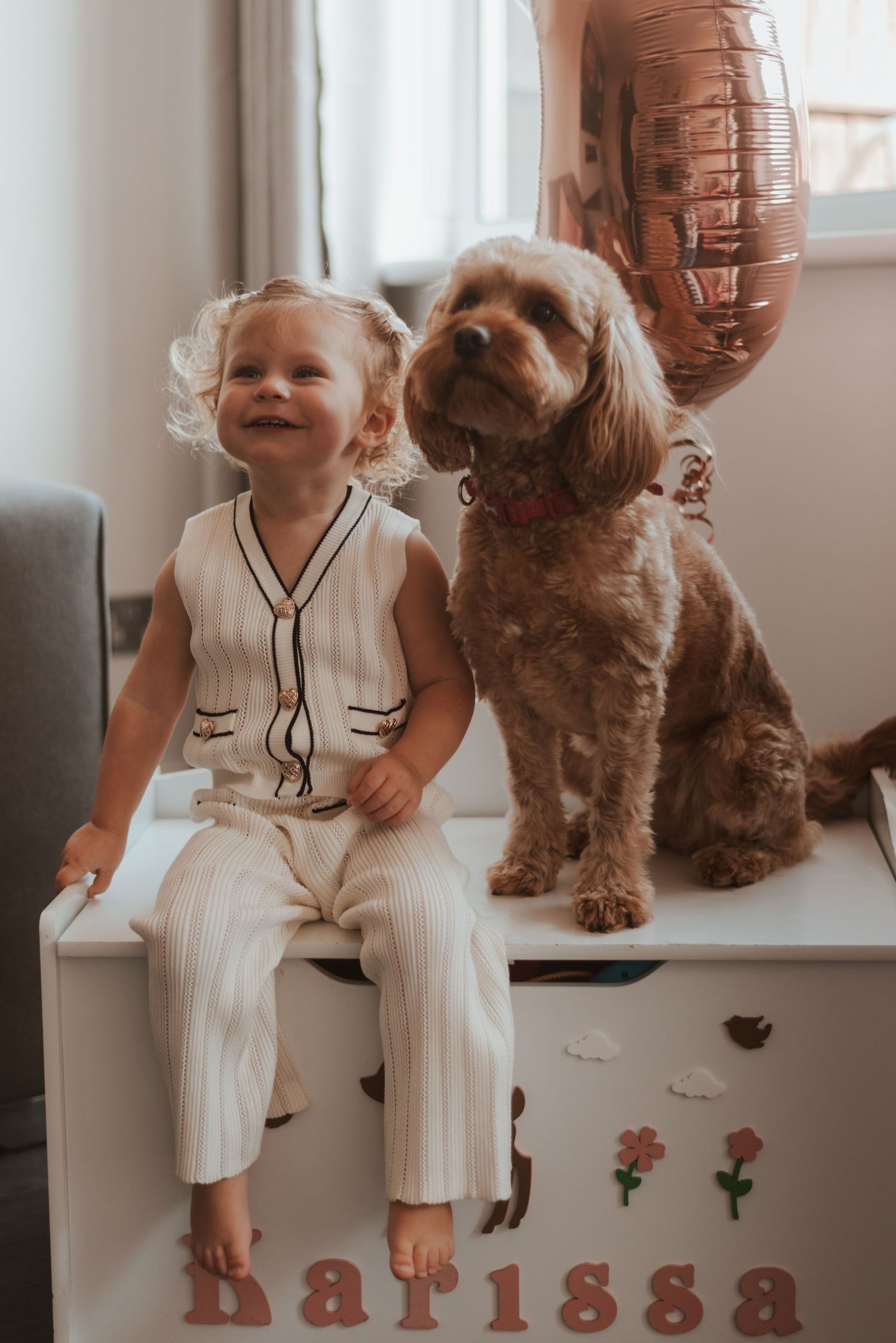 Child in white outfit sitting by window with brown dog and decorative letter display.