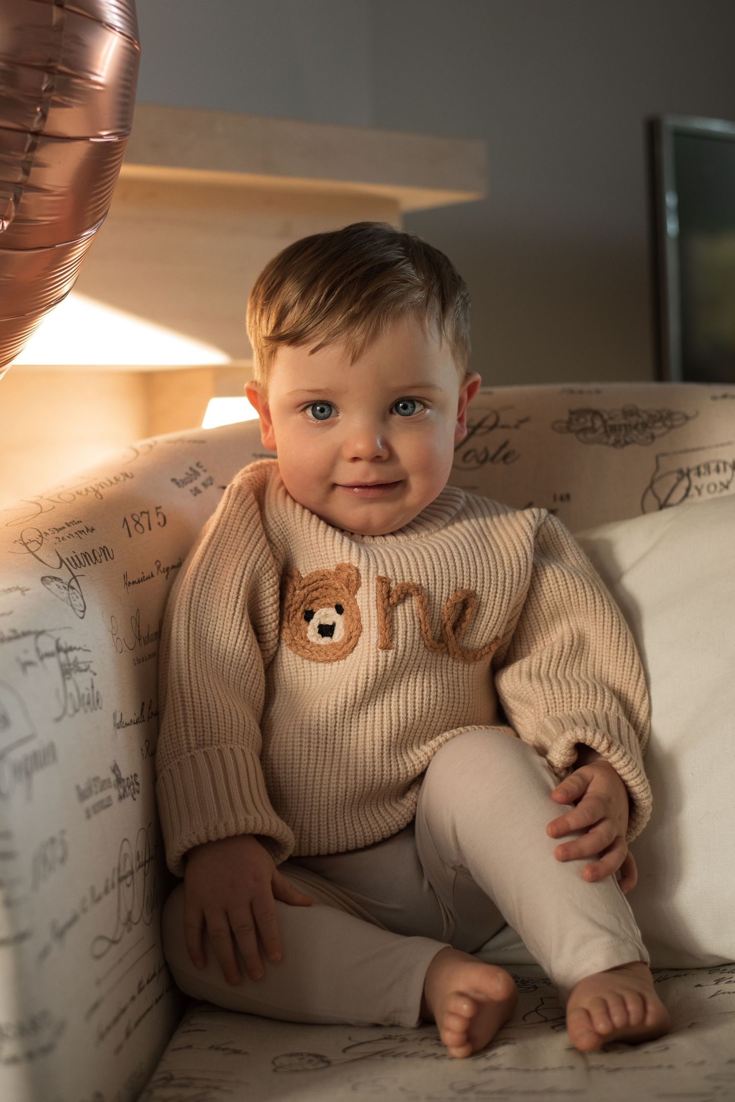 Baby in beige ribbed sweater sitting on couch in warm sunlight.