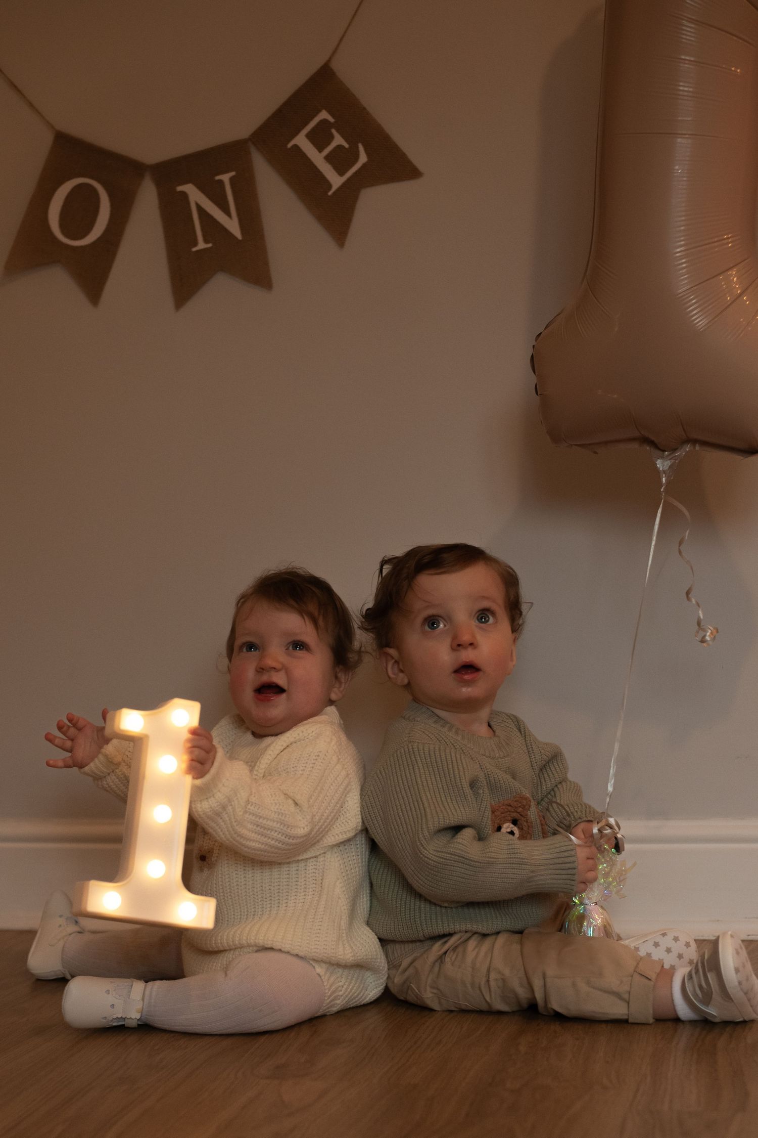 Two toddlers sit by illuminated number one and ONE banner at birthday celebration.