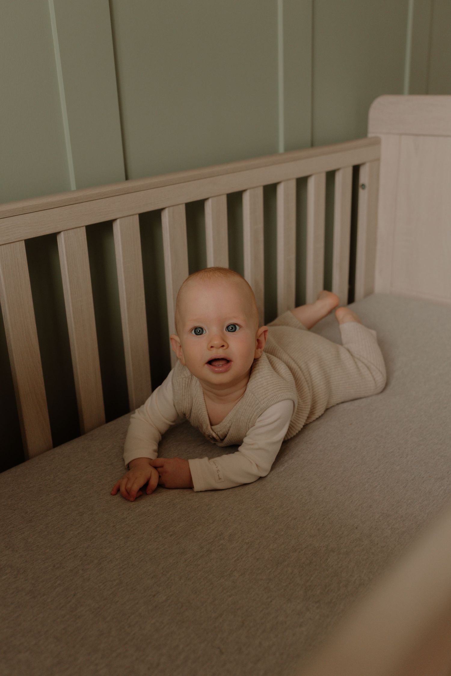 A baby in a cream-colored outfit crawls and plays in a wooden crib with gray bedding.