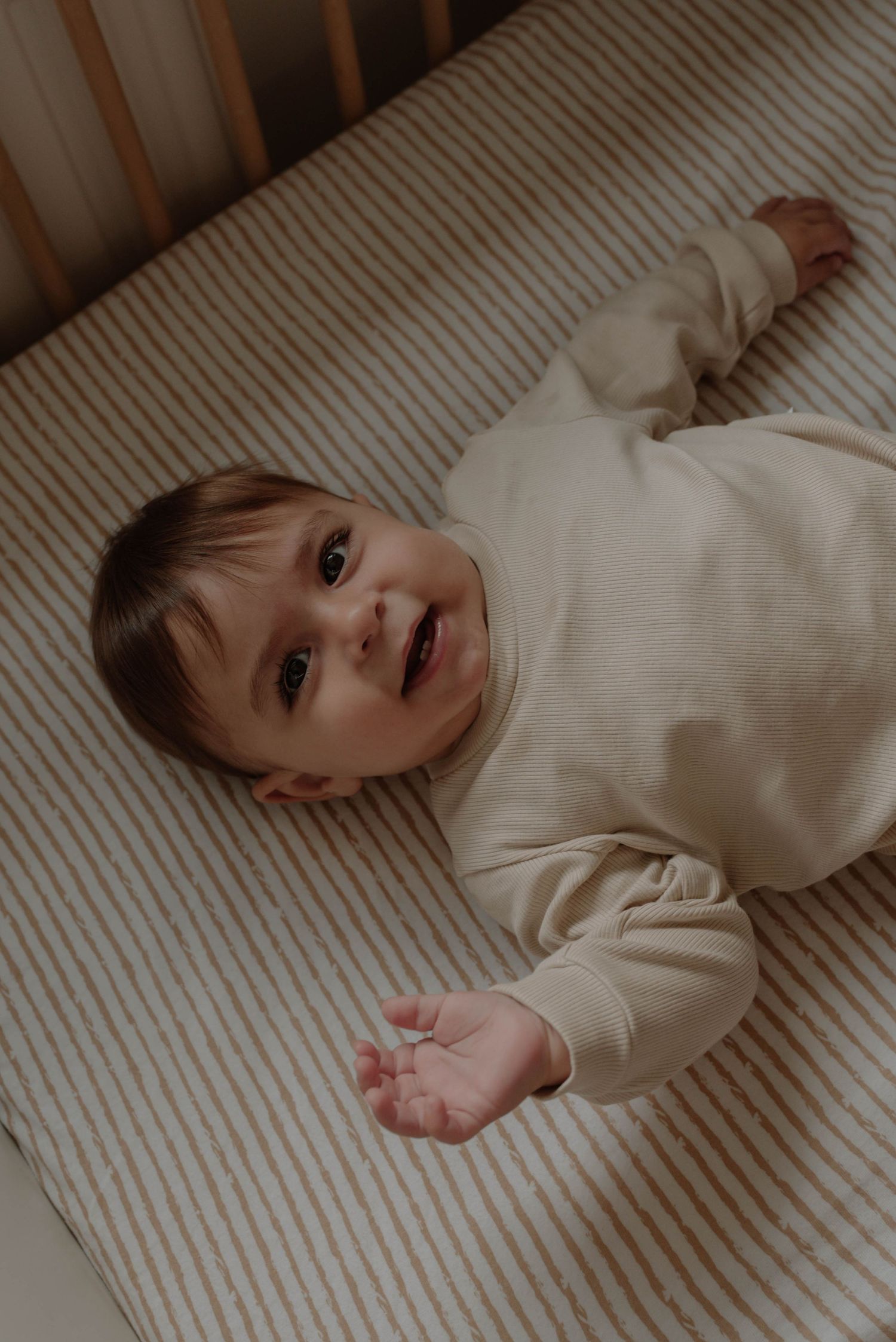 Child in white clothing lying on striped bedding while making playful expressions.