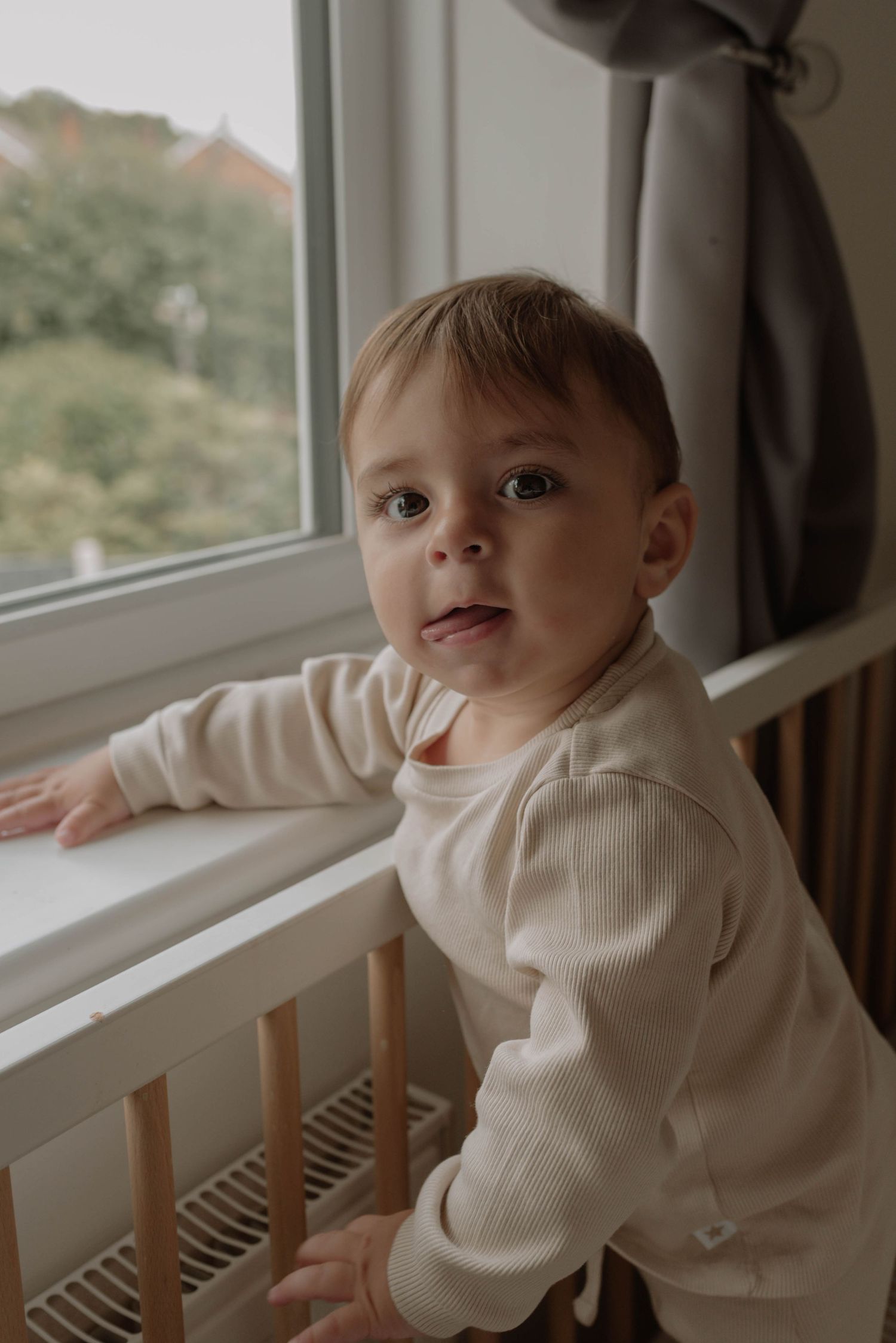 A small child in a cream sweater looks out a window while standing in a wooden crib.