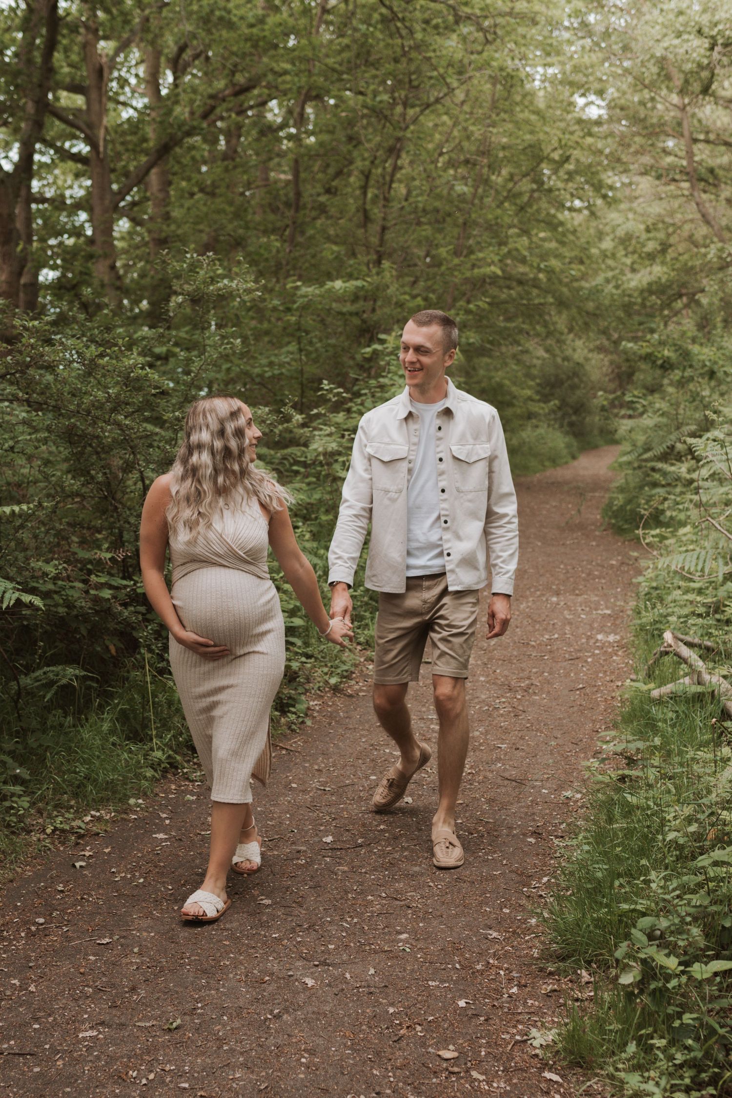 A couple walks together on a scenic dirt trail through lush green woodland on a sunny afternoon.