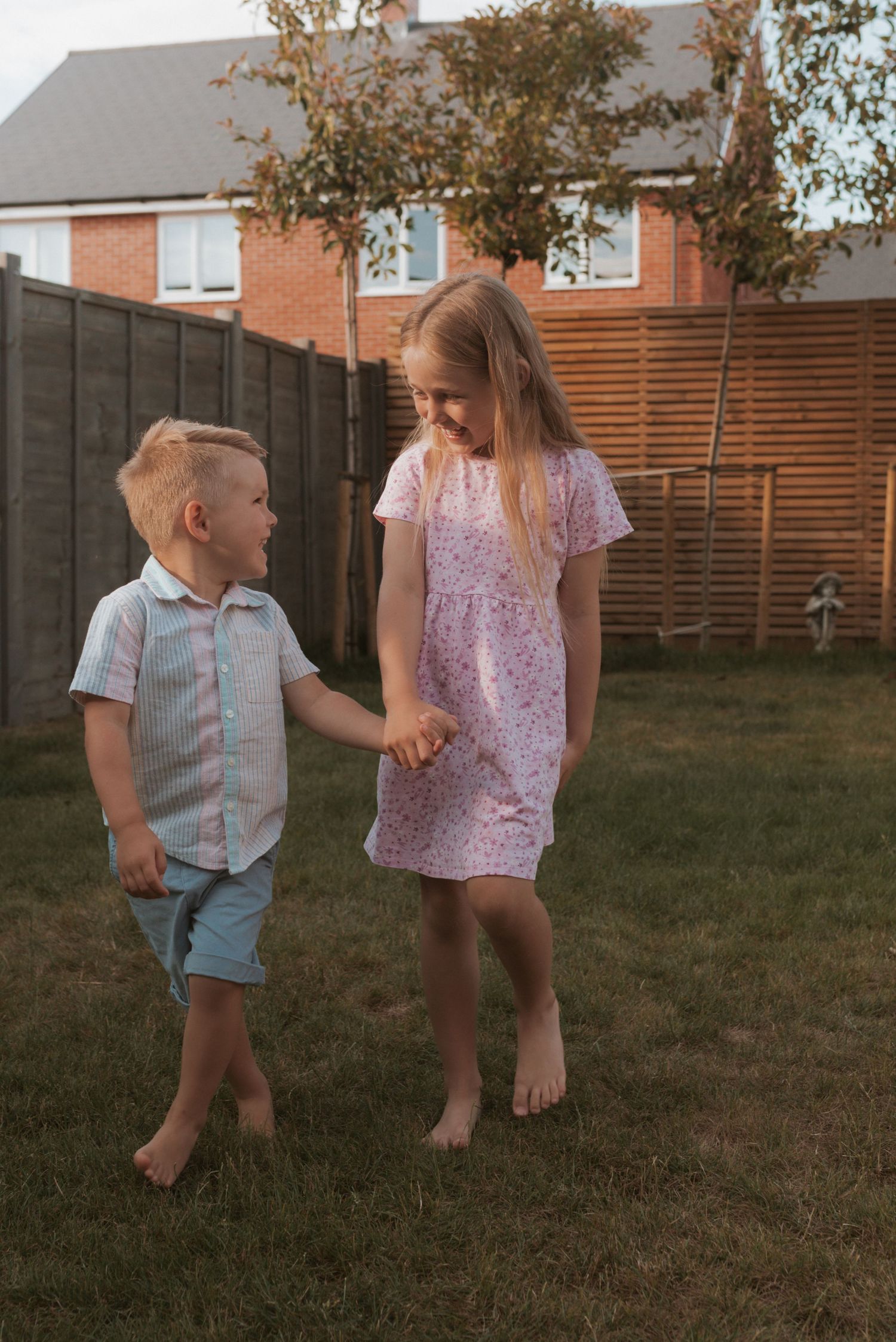 Two children in summer outfits holding hands and smiling in a backyard at sunset.