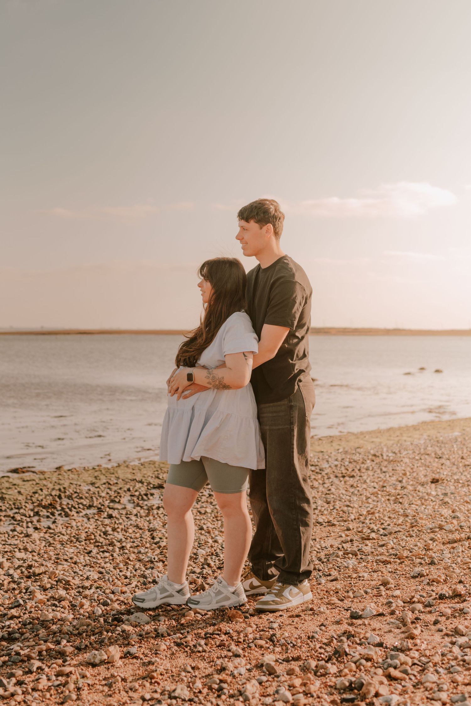 A couple shares a romantic moment on a pebble beach at sunset during their maternity shoot.