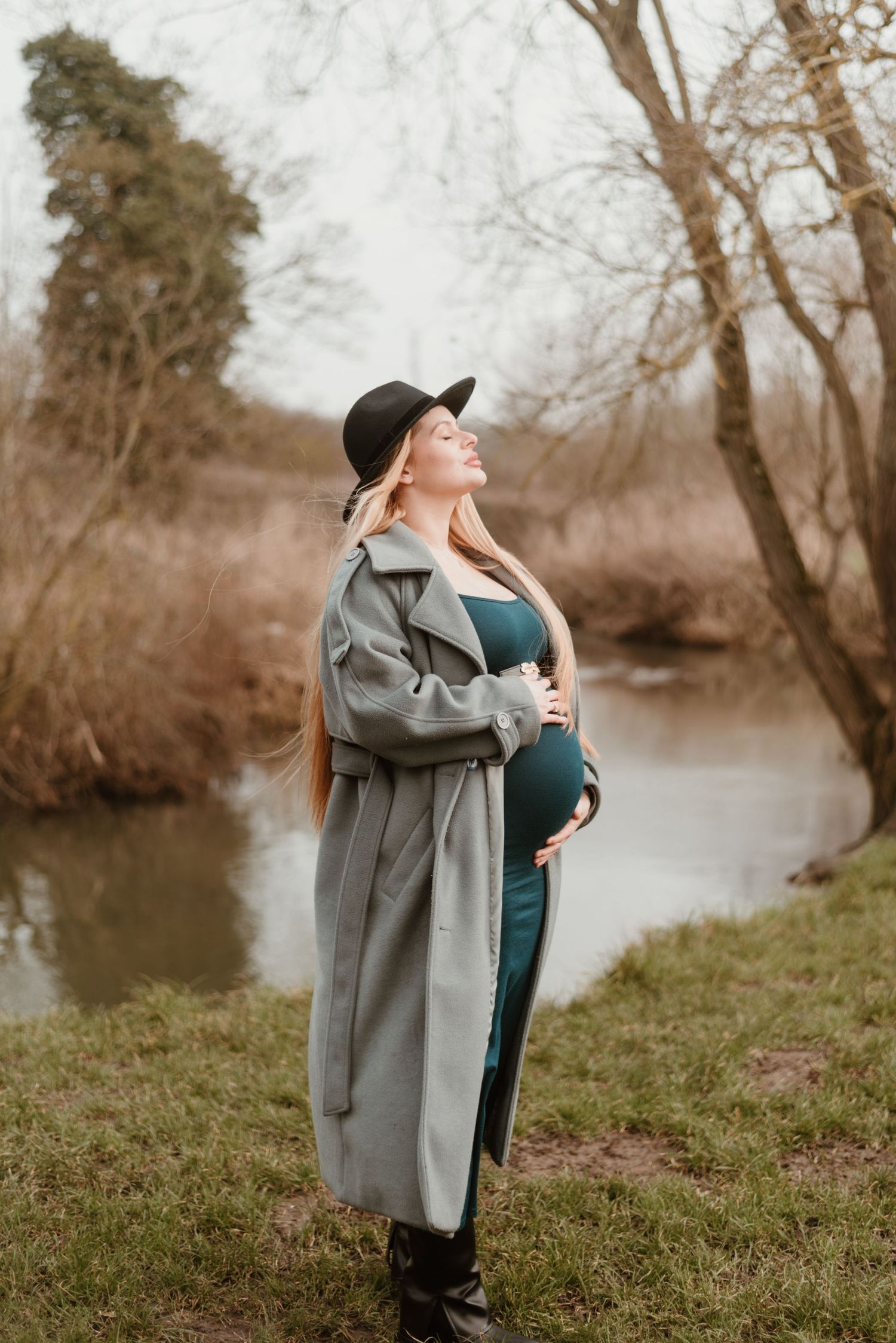 A stylish maternity photo by a river featuring a long gray coat and black hat in autumn scenery.