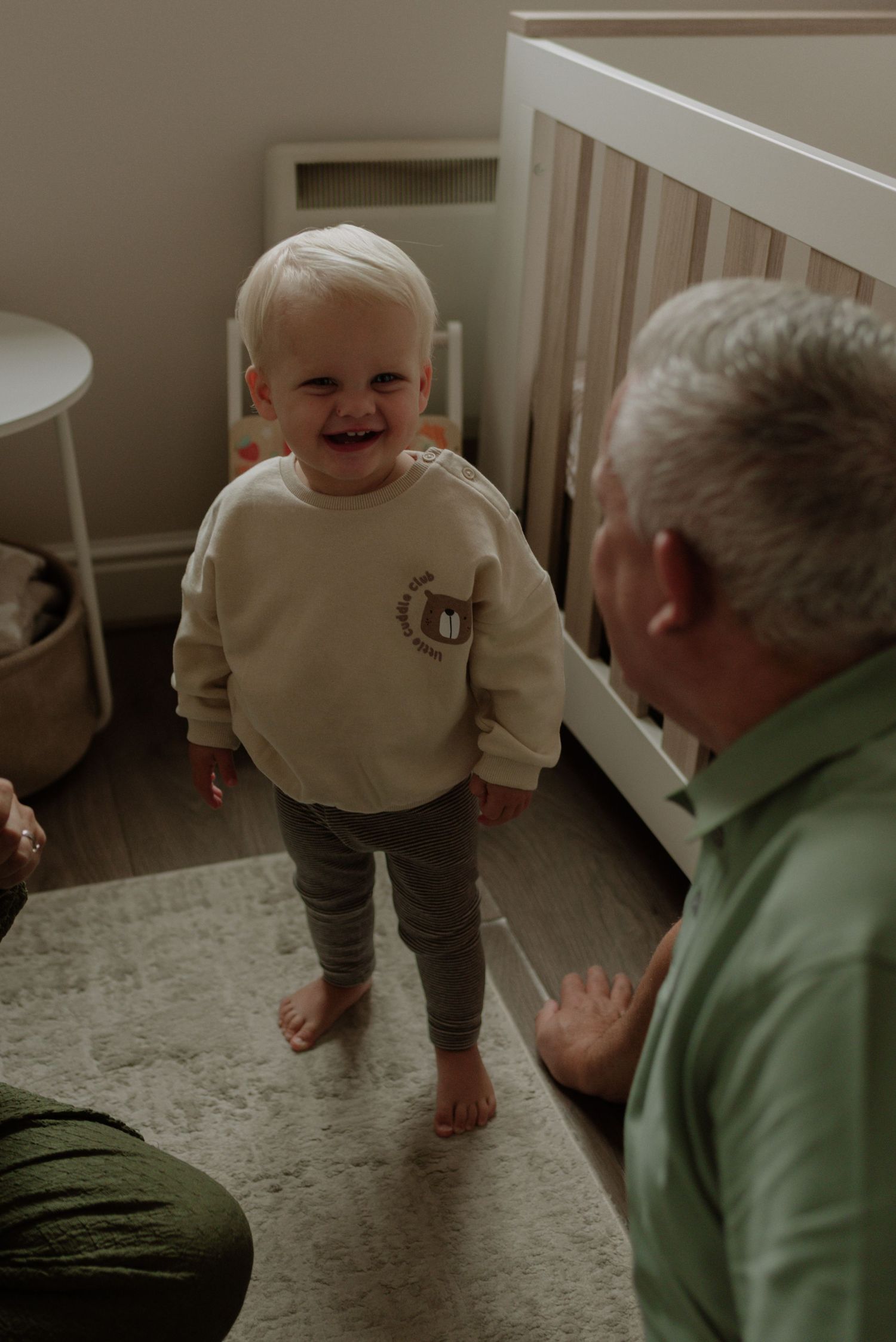 A small child in a beige sweater and gray pants stands barefoot on carpet while interacting with someone in green.