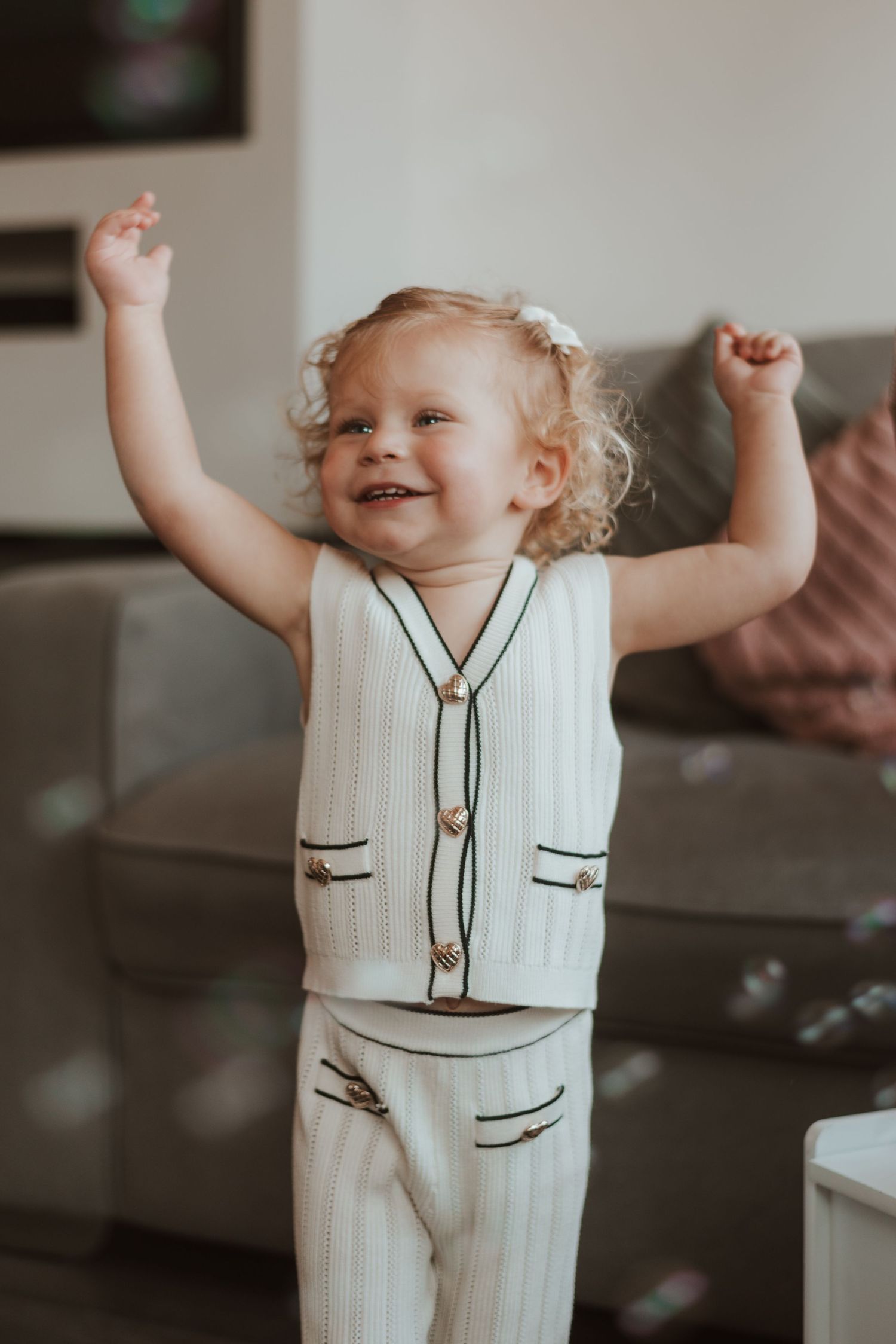 A joyful toddler raises their arms while wearing a white ribbed outfit.