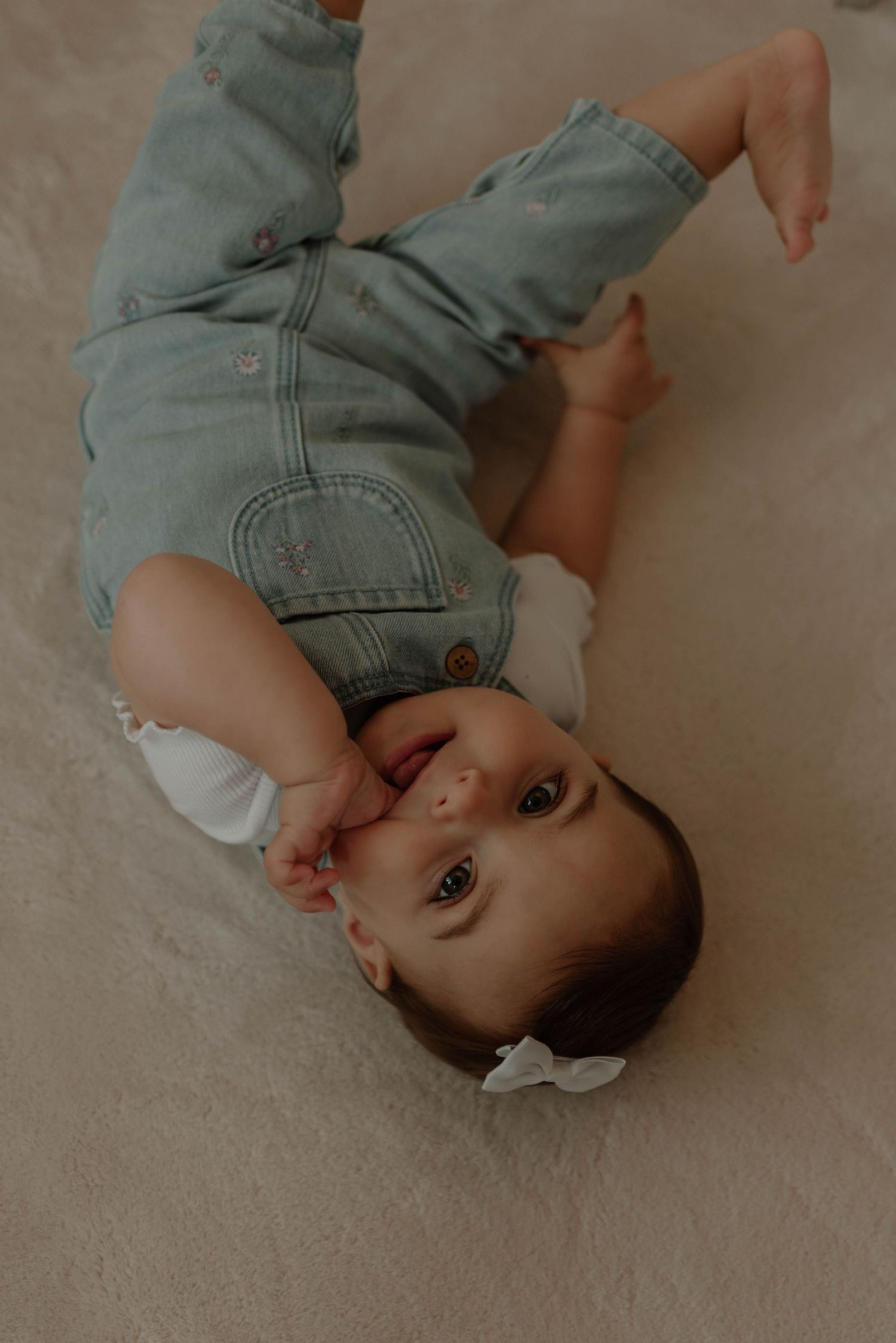 A baby in denim overalls lies on a beige carpet while looking up at the camera.
