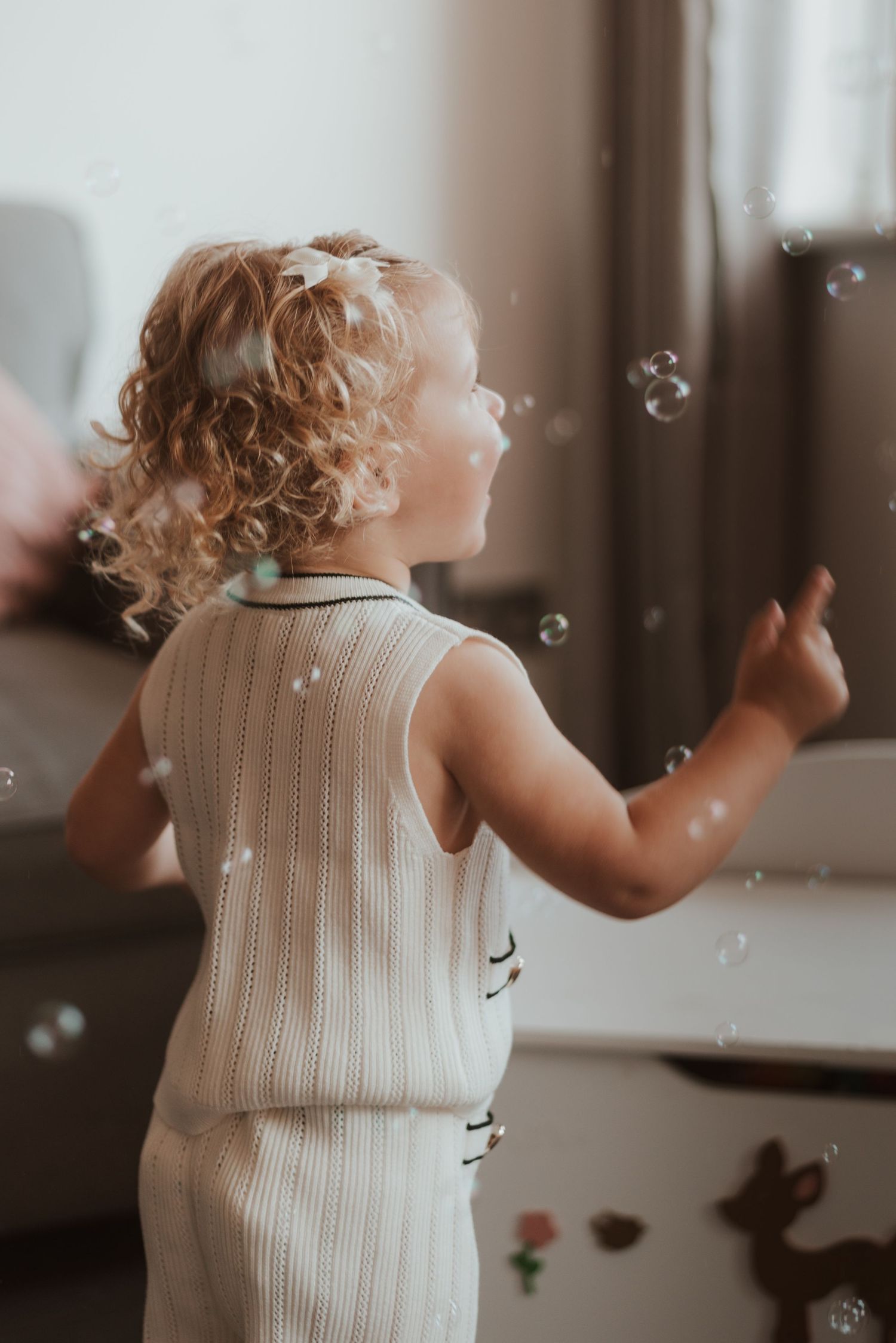Small child in white dress watches floating soap bubbles with wonder in soft, warm lighting by window.