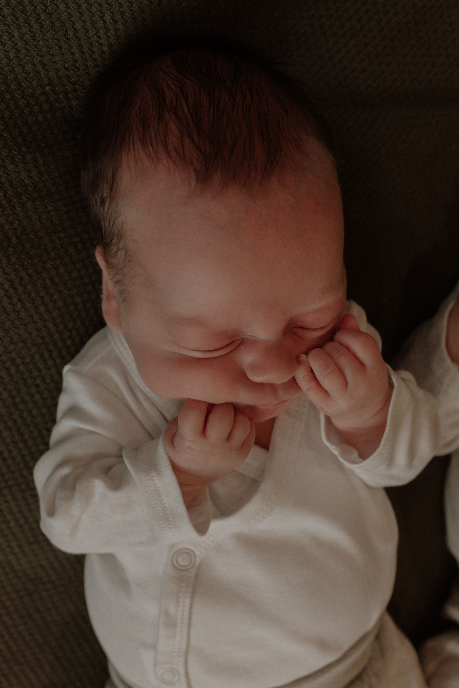 A sleeping baby in a white onesie rubs their eyes while resting on a dark textured surface.