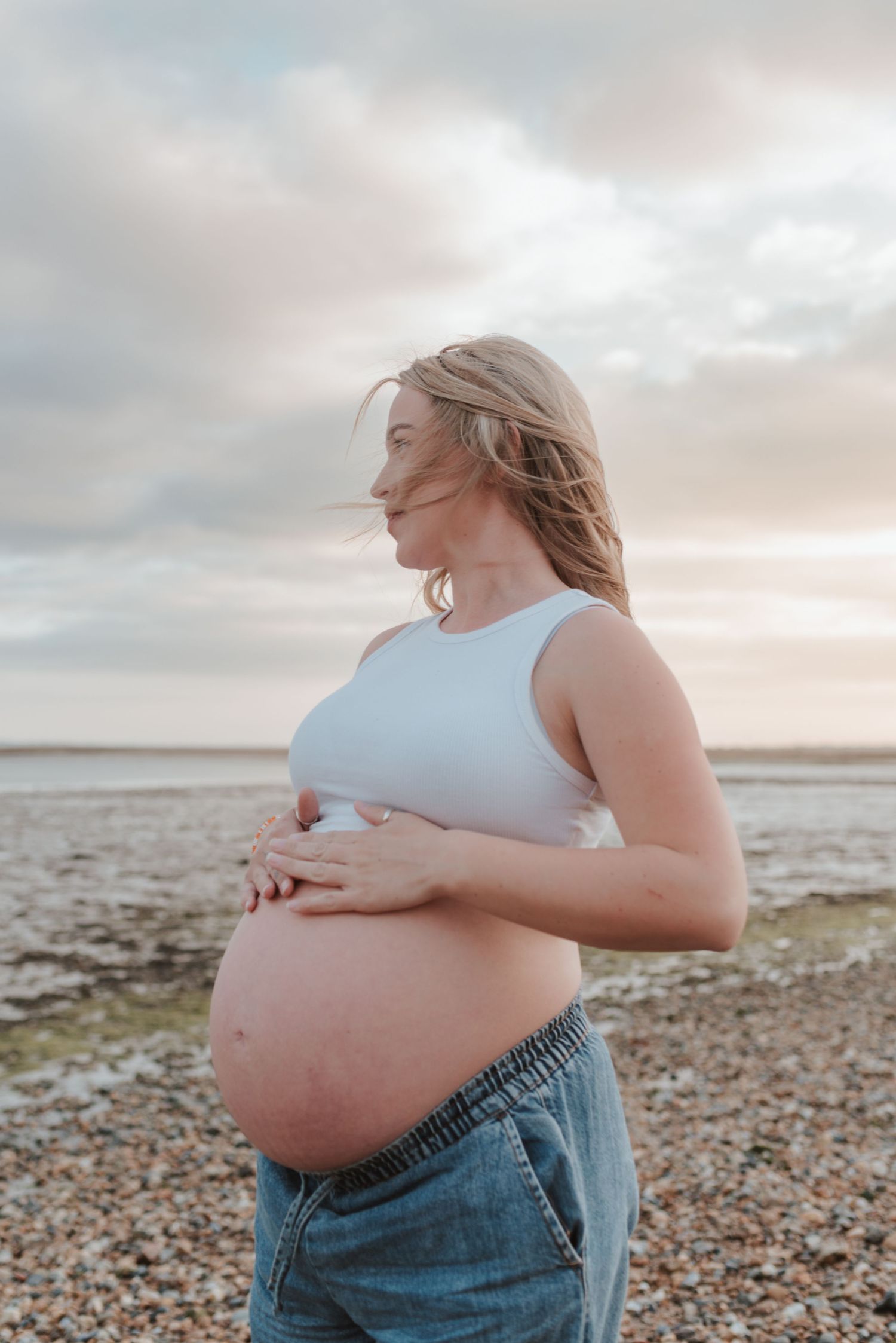 Pregnant woman in white tank top standing on a rocky beach at sunset.