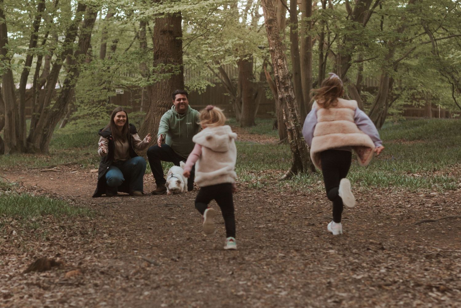 A family playing together in a wooded area, jumping and moving across a natural dirt ground.