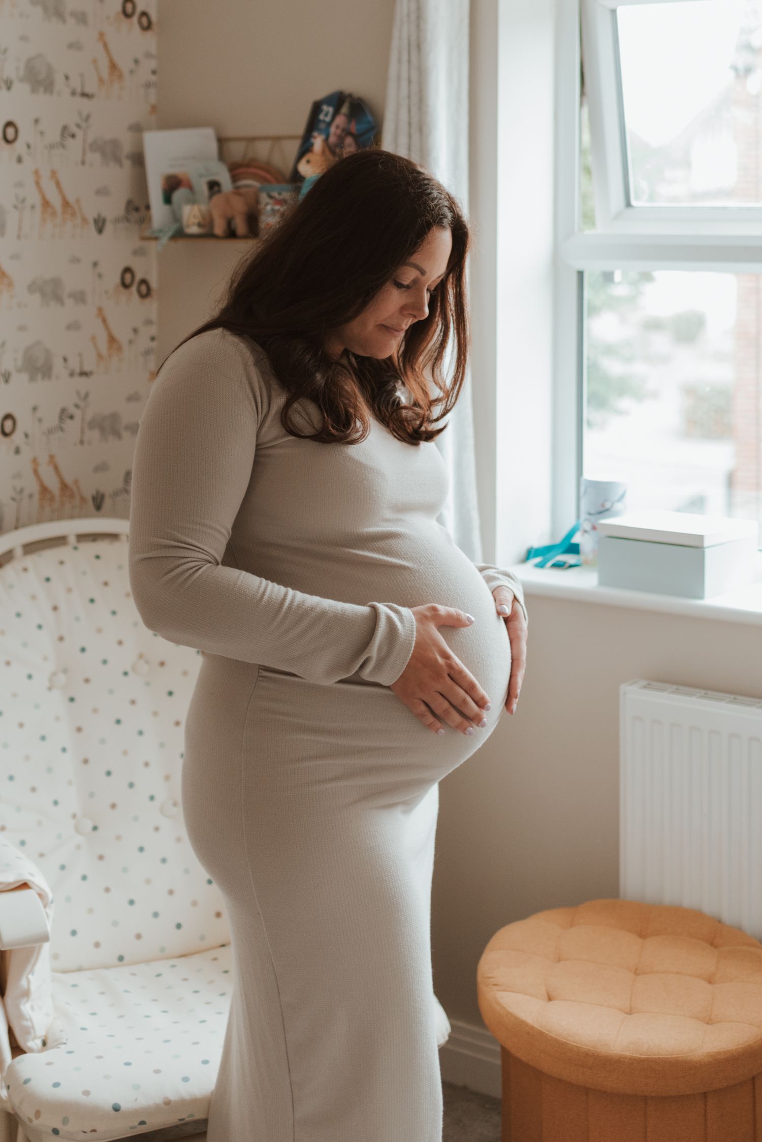 Side view of pregnant woman in grey dress standing by window in nursery.