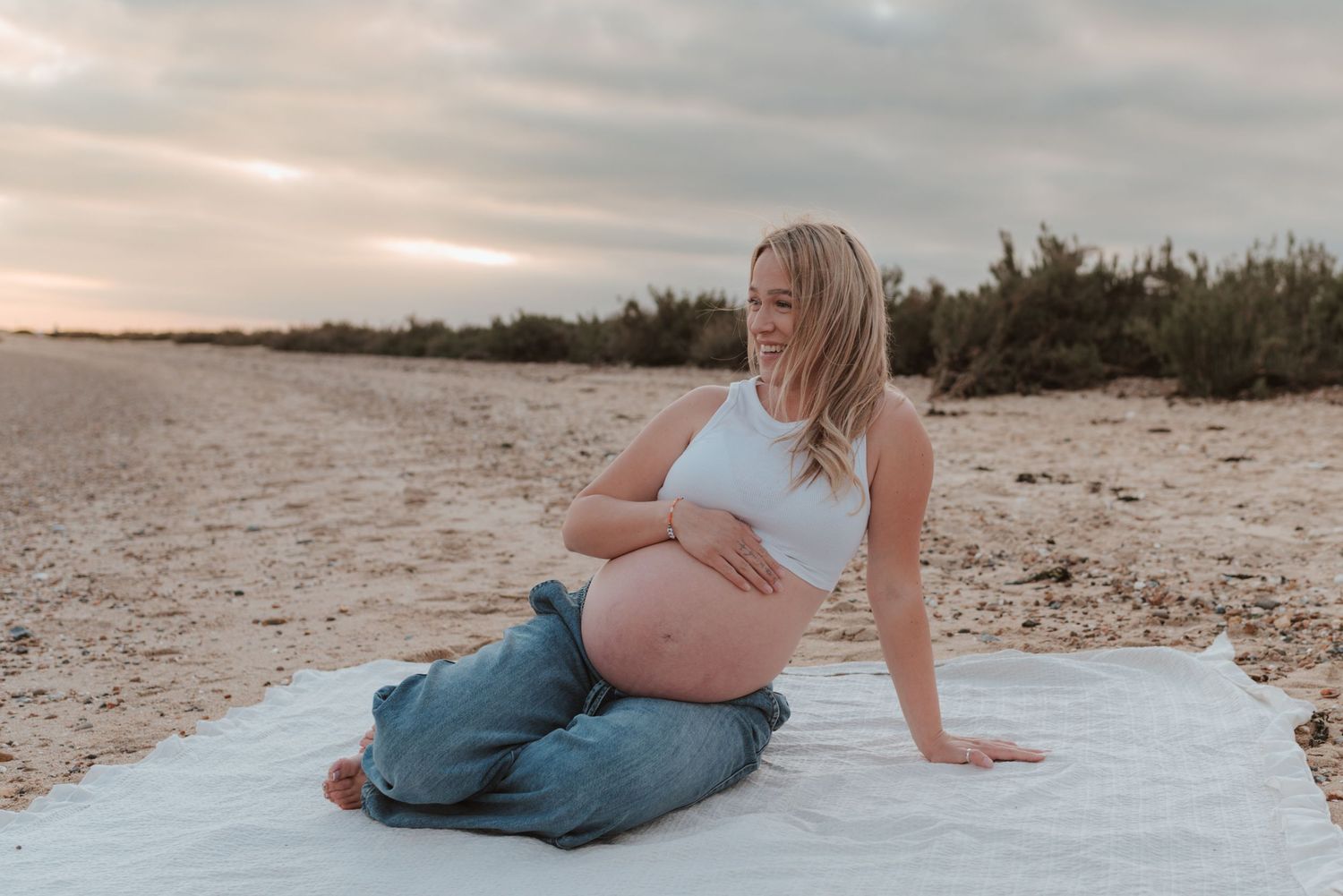 A tranquil maternity photo on a sandy beach at sunset with soft lighting and a white blanket backdrop.