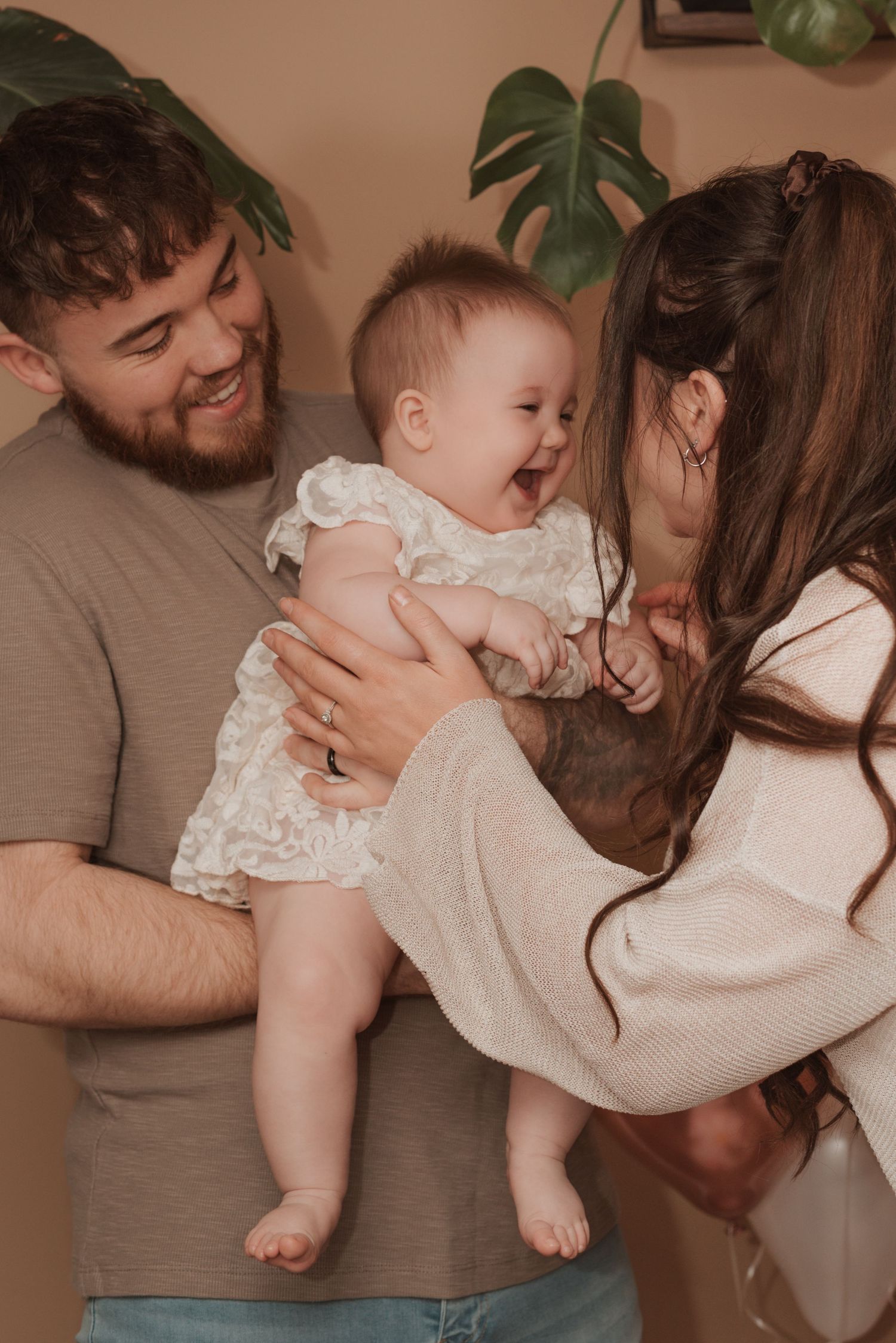 A joyful family moment as parents share laughter with their baby wearing a white lace dress.