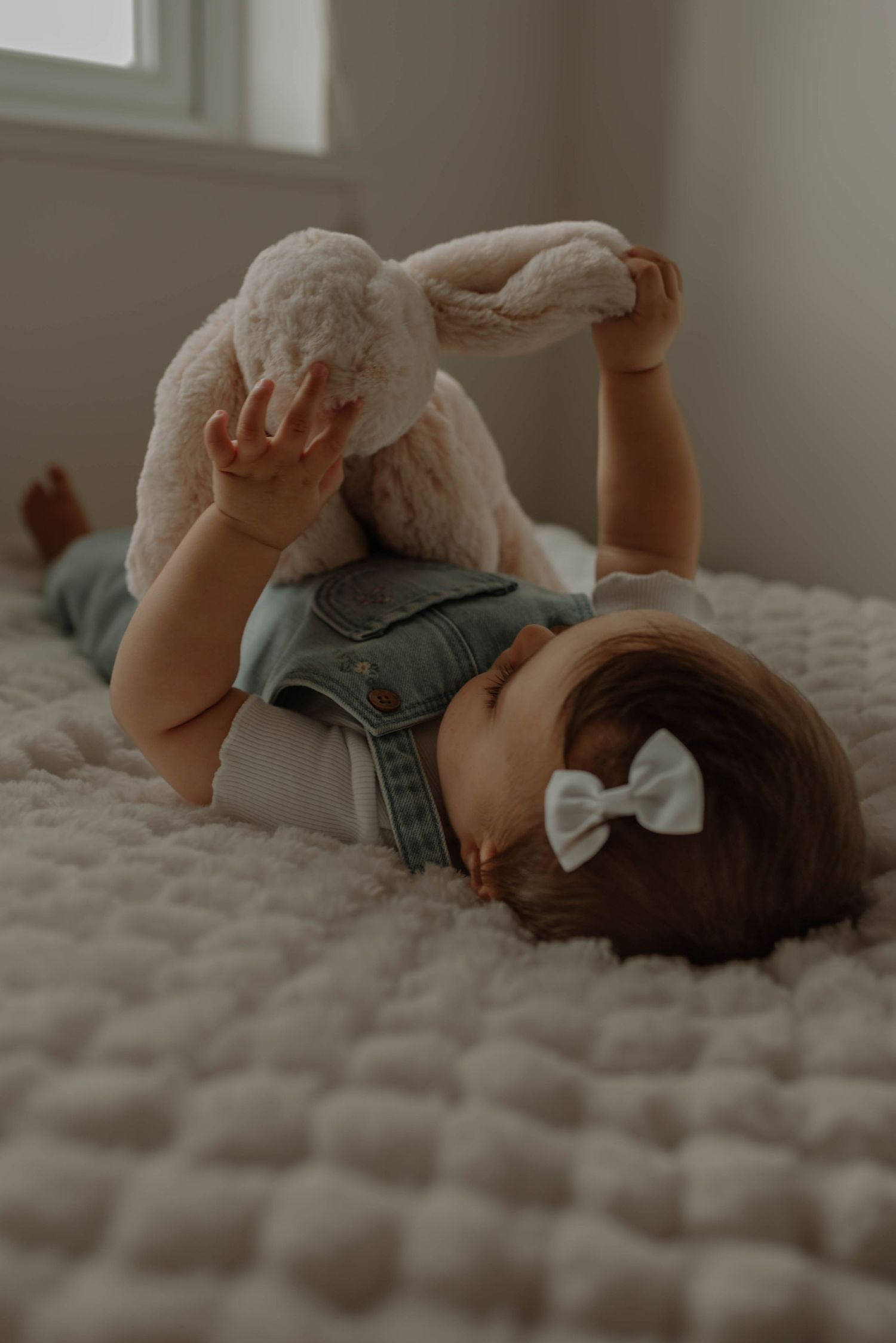 Baby in denim overalls rolls playfully on textured white bedding in a series of candid poses.