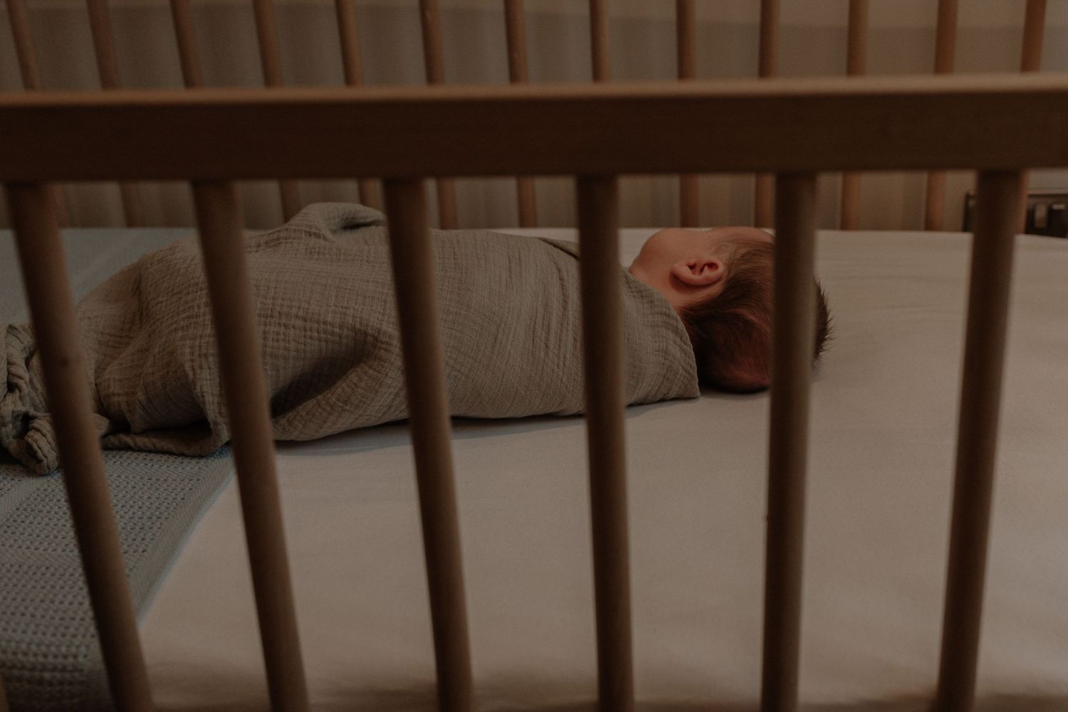 View through wooden crib slats showing cozy bedding and a stuffed animal in dim lighting.