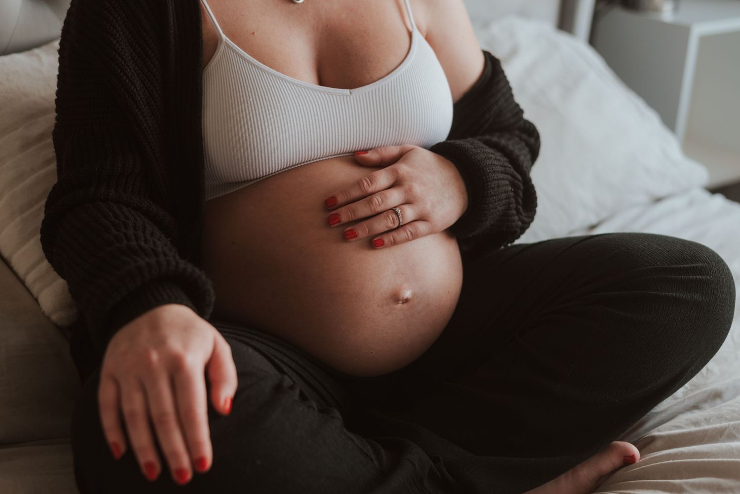 Pregnant woman in black sweater holding her baby bump while sitting comfortably on a bed.