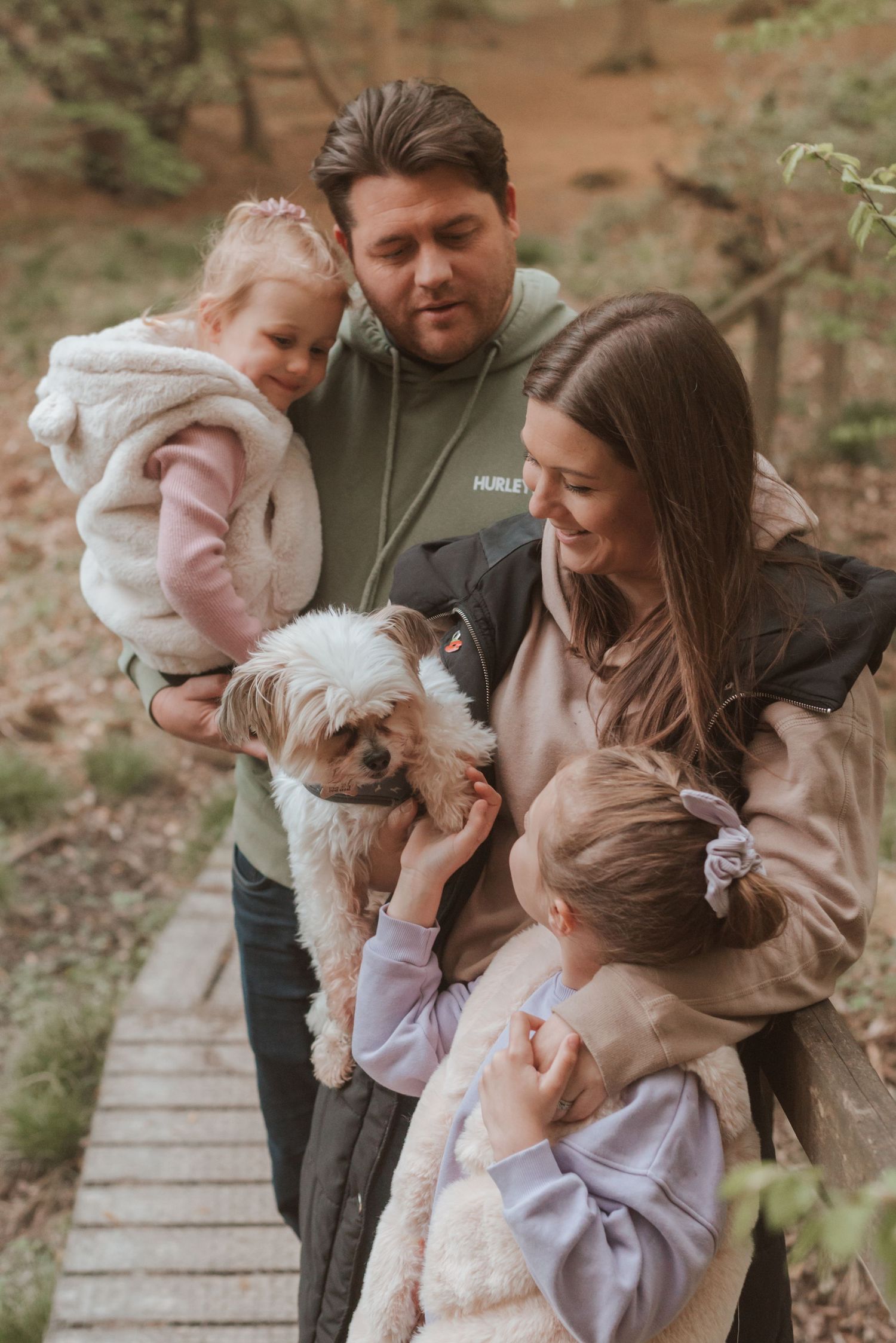 A family and their small fluffy dog share a moment together on a wooden boardwalk in nature.