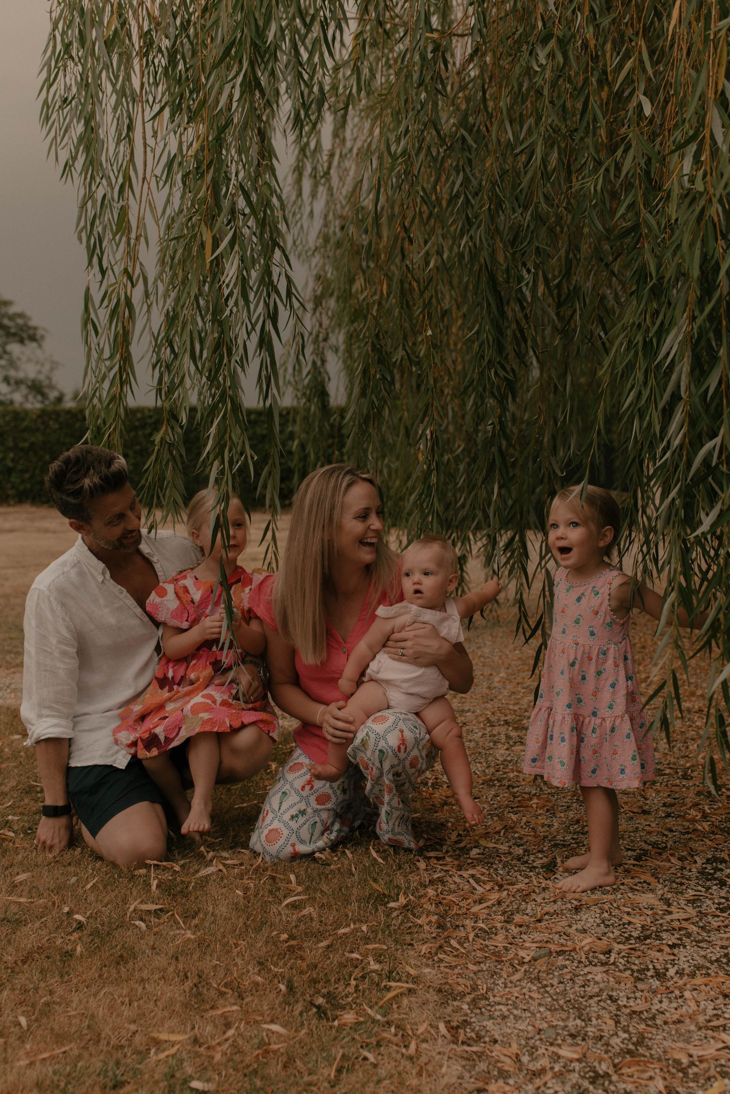 A family in coordinated coral and white outfits sits together under a drooping willow tree.