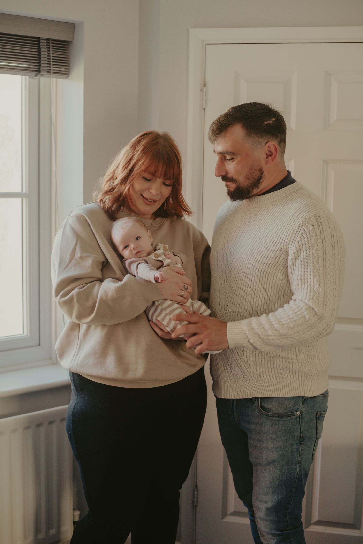 Family wearing matching cream sweaters sharing intimate moments with their baby in a bright room.