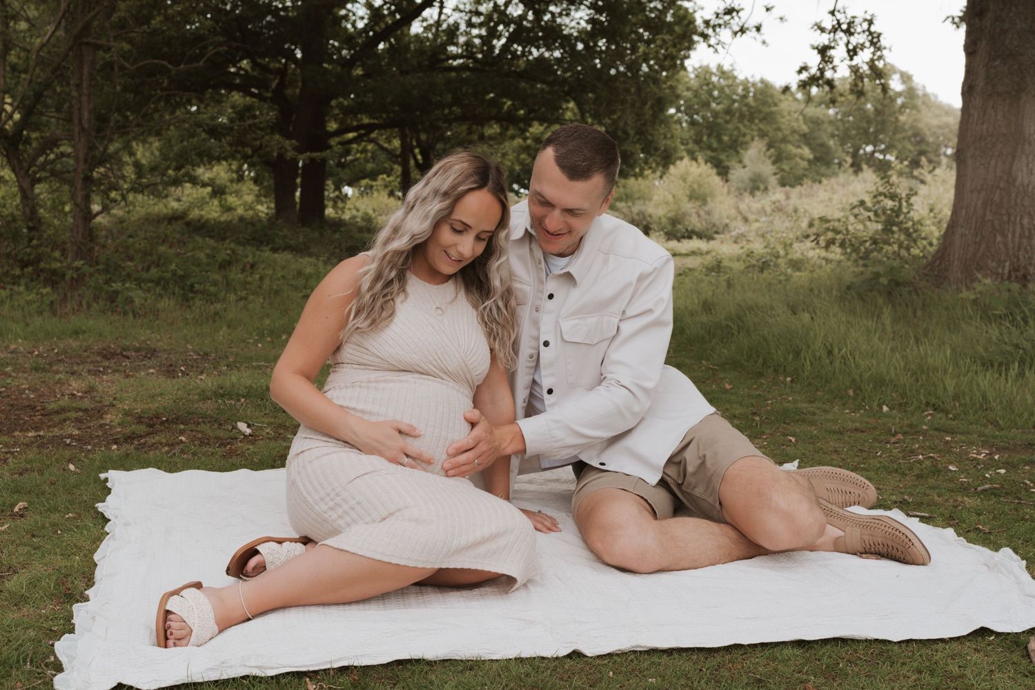 A couple in white outfits sharing a tender moment on a blanket in a sunny park setting.