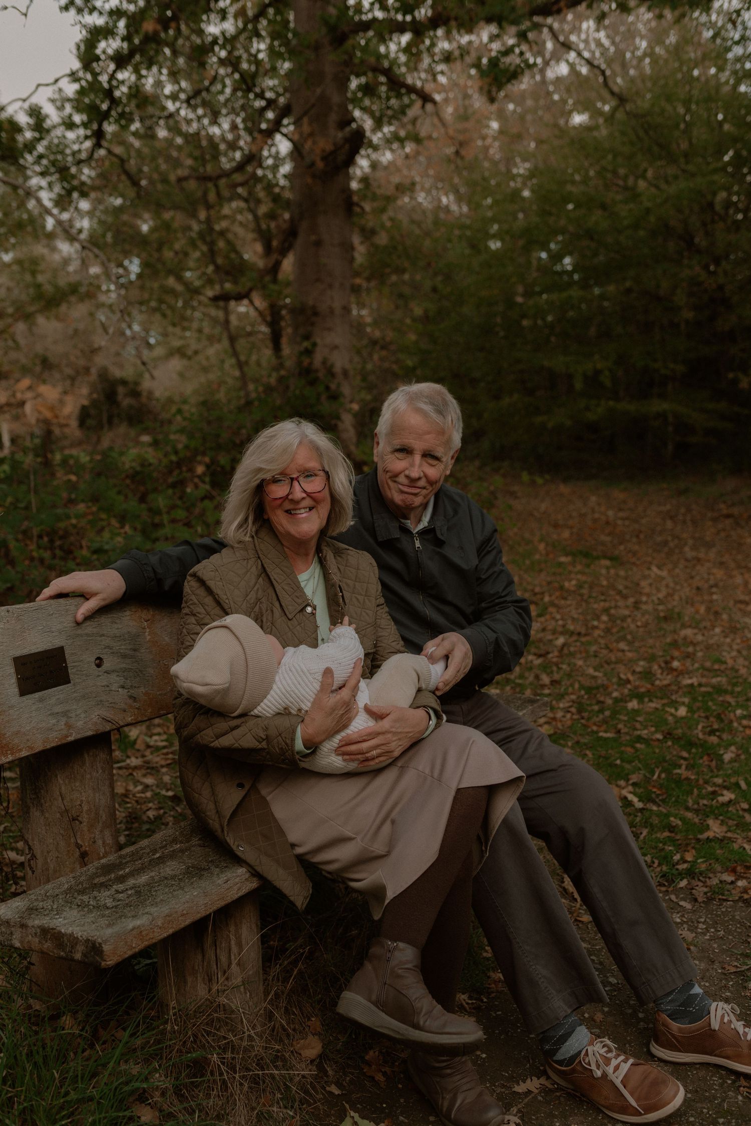 A couple sits together on a rustic wooden bench surrounded by autumn leaves in a forest setting.