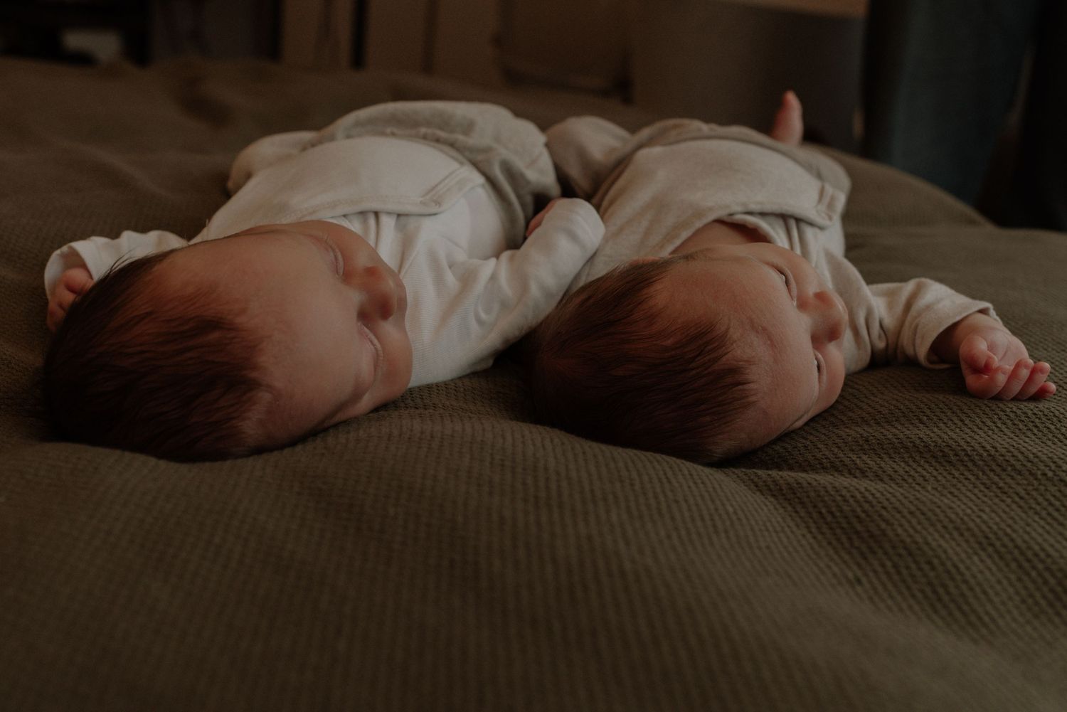 Two sleeping infants in white onesies lying side by side on a green textured surface.