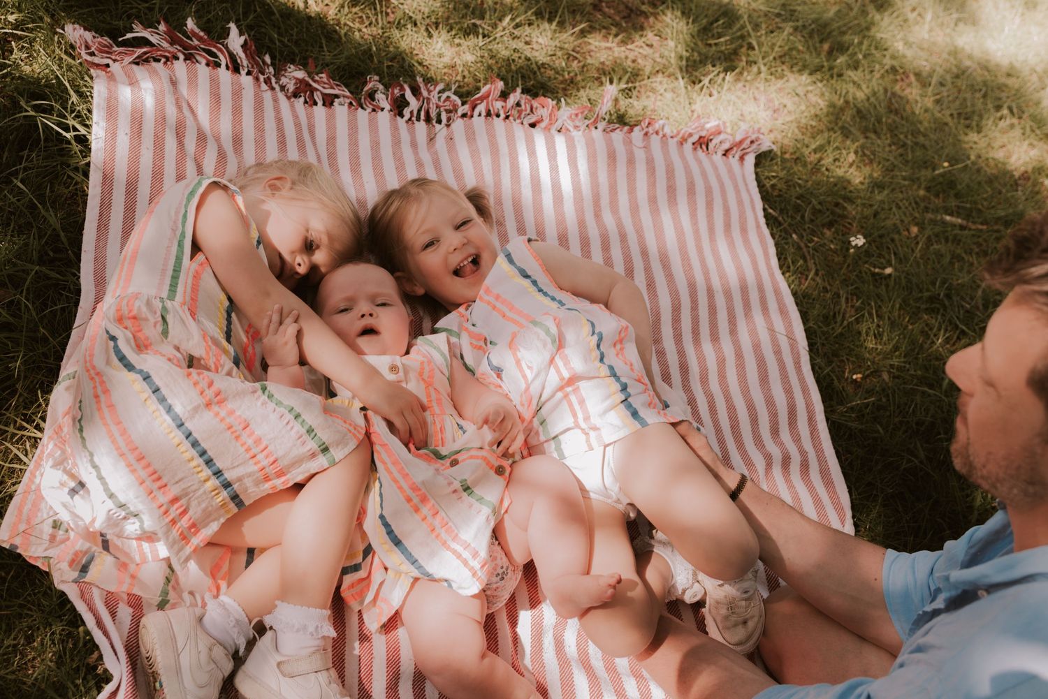 Three young children laying on a pink striped blanket outdoors while being entertained.