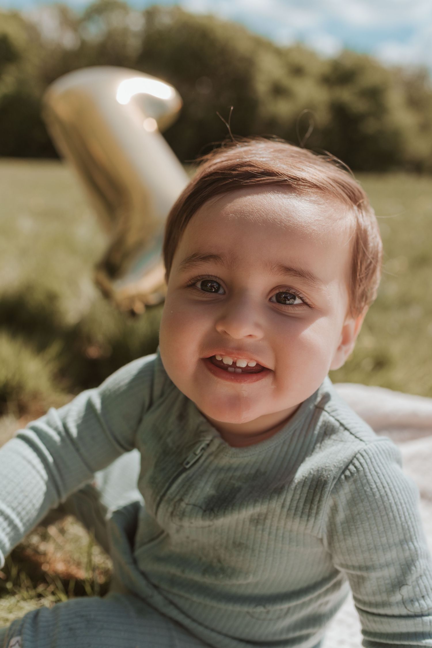 A smiling toddler wearing a mint green sweater sits on grass in warm sunlight.