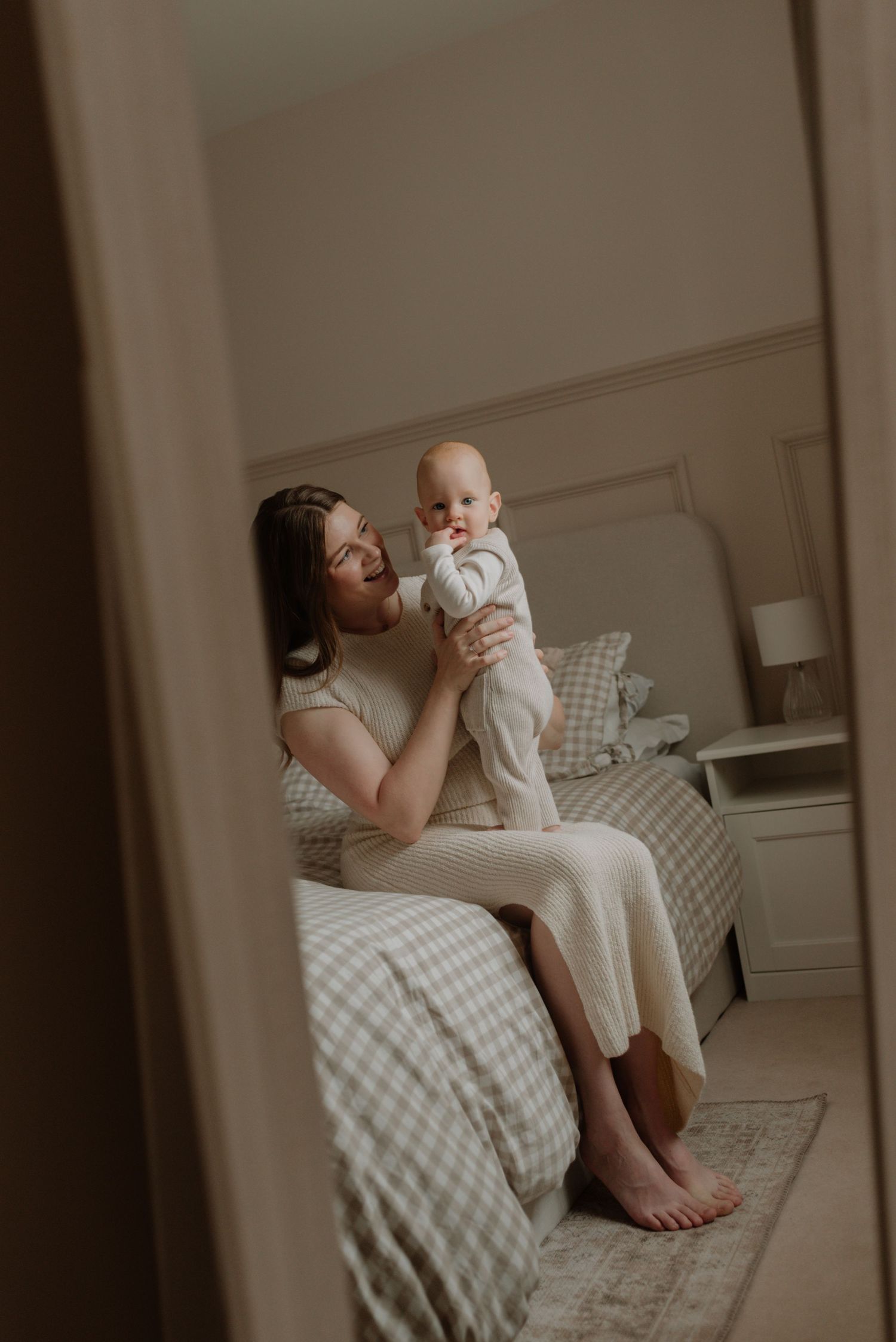 A parent and baby share a tender moment on a bed with plaid bedding and neutral decor in a softly lit bedroom.