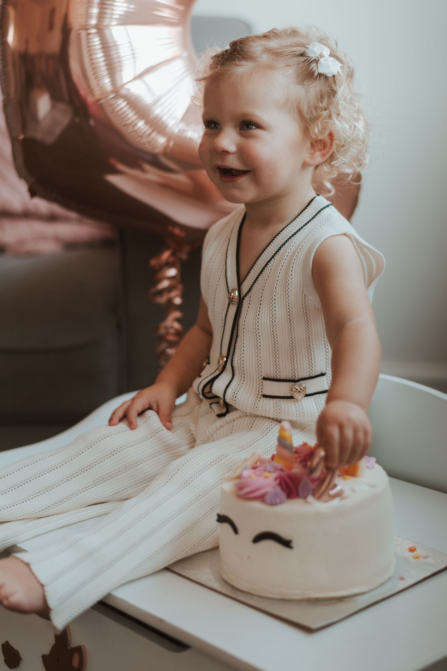 Baby in white dress sitting on white bedding while reaching for unseen object in a softly lit room.