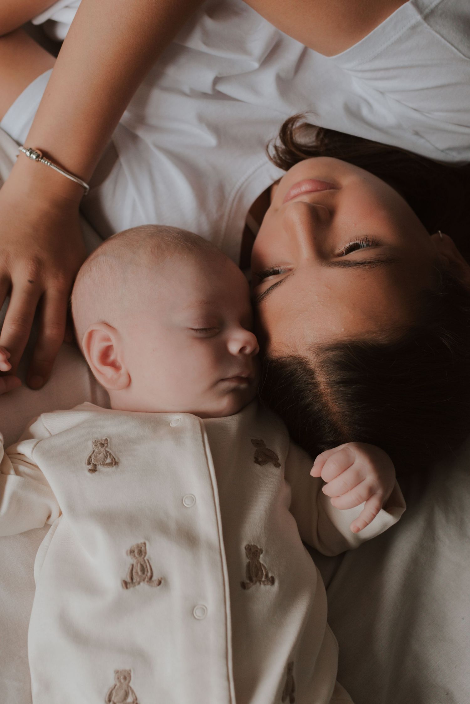 A sleeping baby in a teddy bear print onesie rests peacefully next to someone in white.