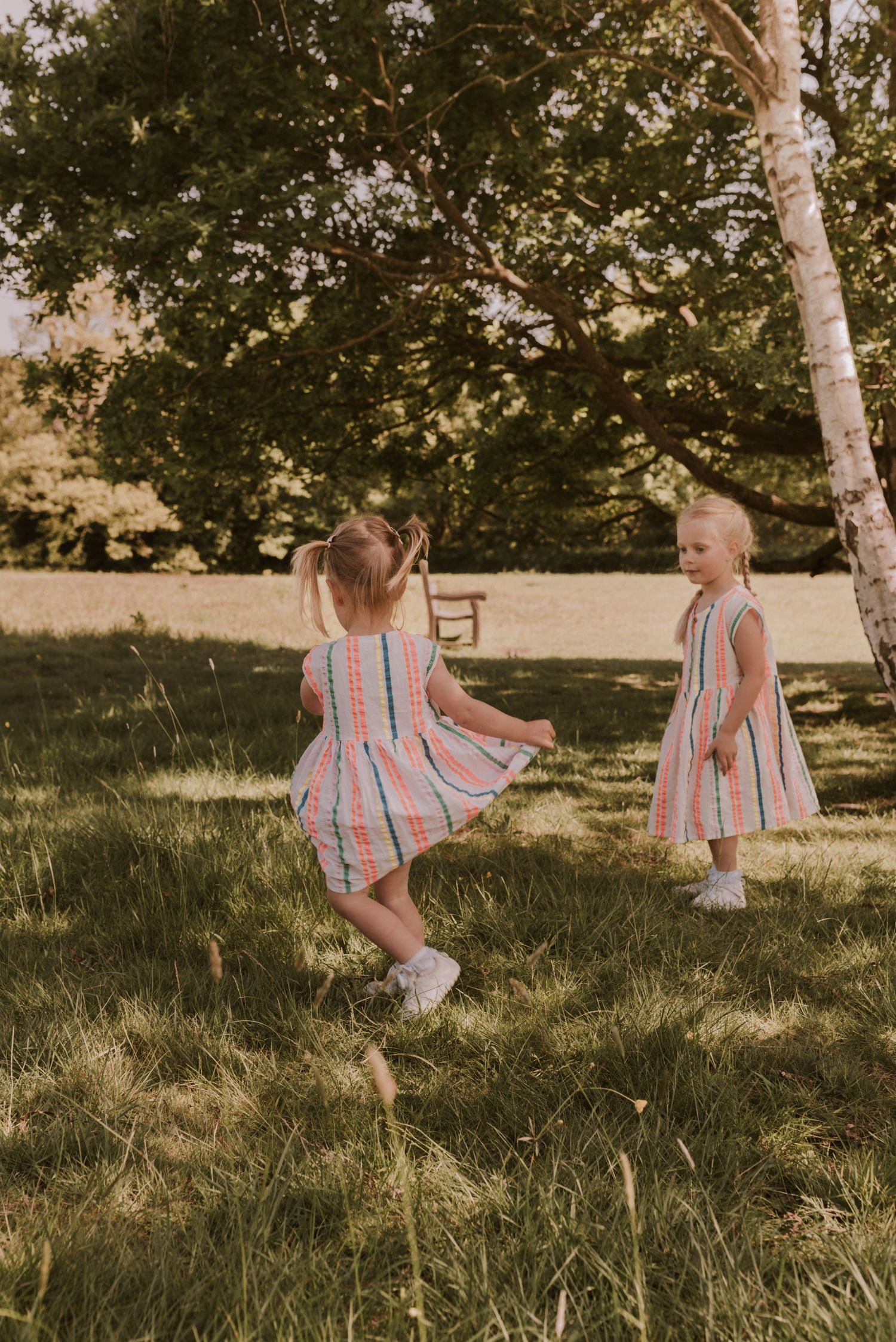 Two children in matching striped dresses playing and twirling in a sunny garden.