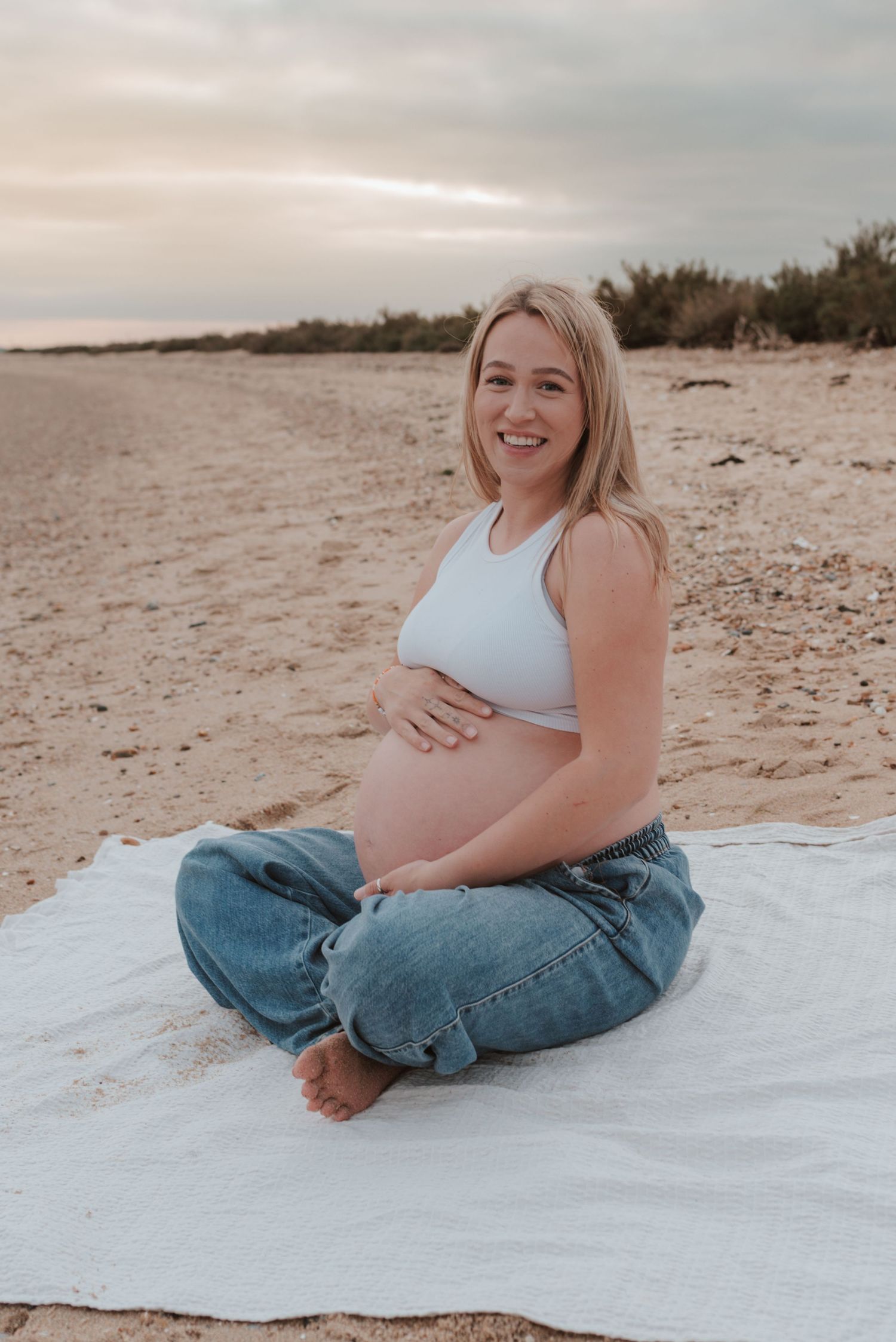 Maternity photoshoot at the beach with white blanket and casual denim at sunset.