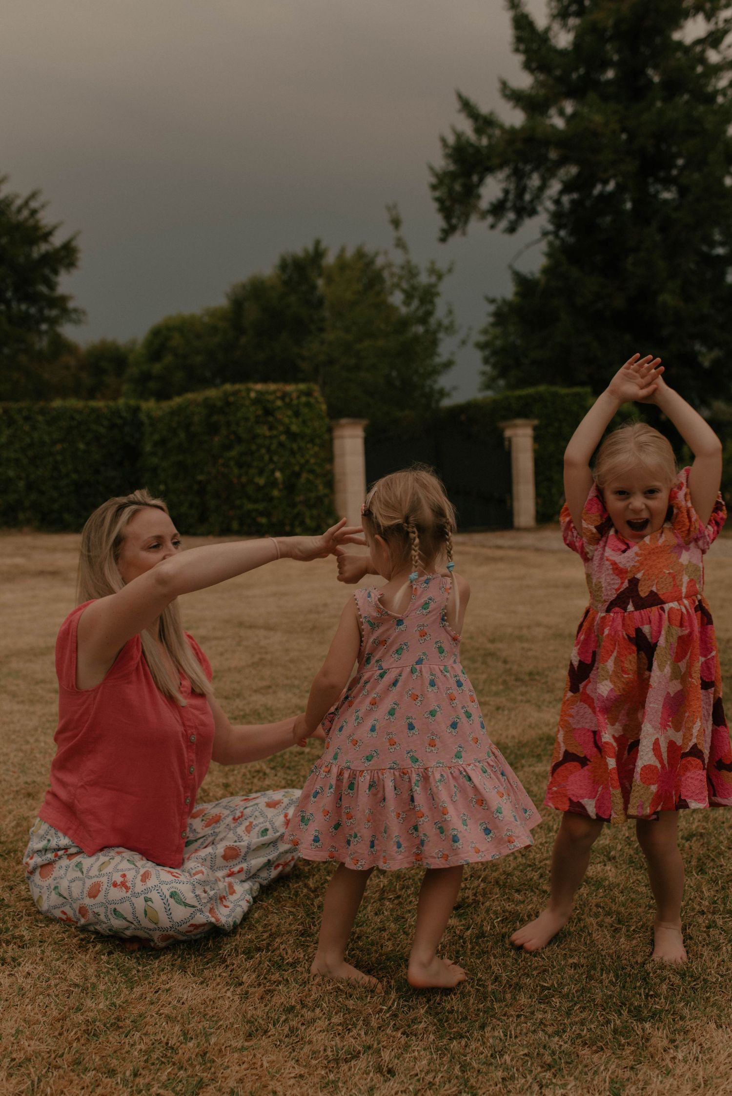 Three people in summer dresses dance and play together on a lawn with hedges and pillars in the background at dusk.