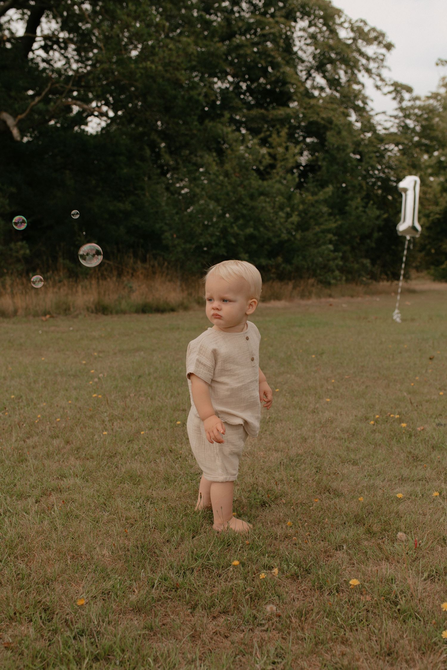 A small child in beige clothing chases bubbles while playing outdoors in a grassy field on a sunny day.