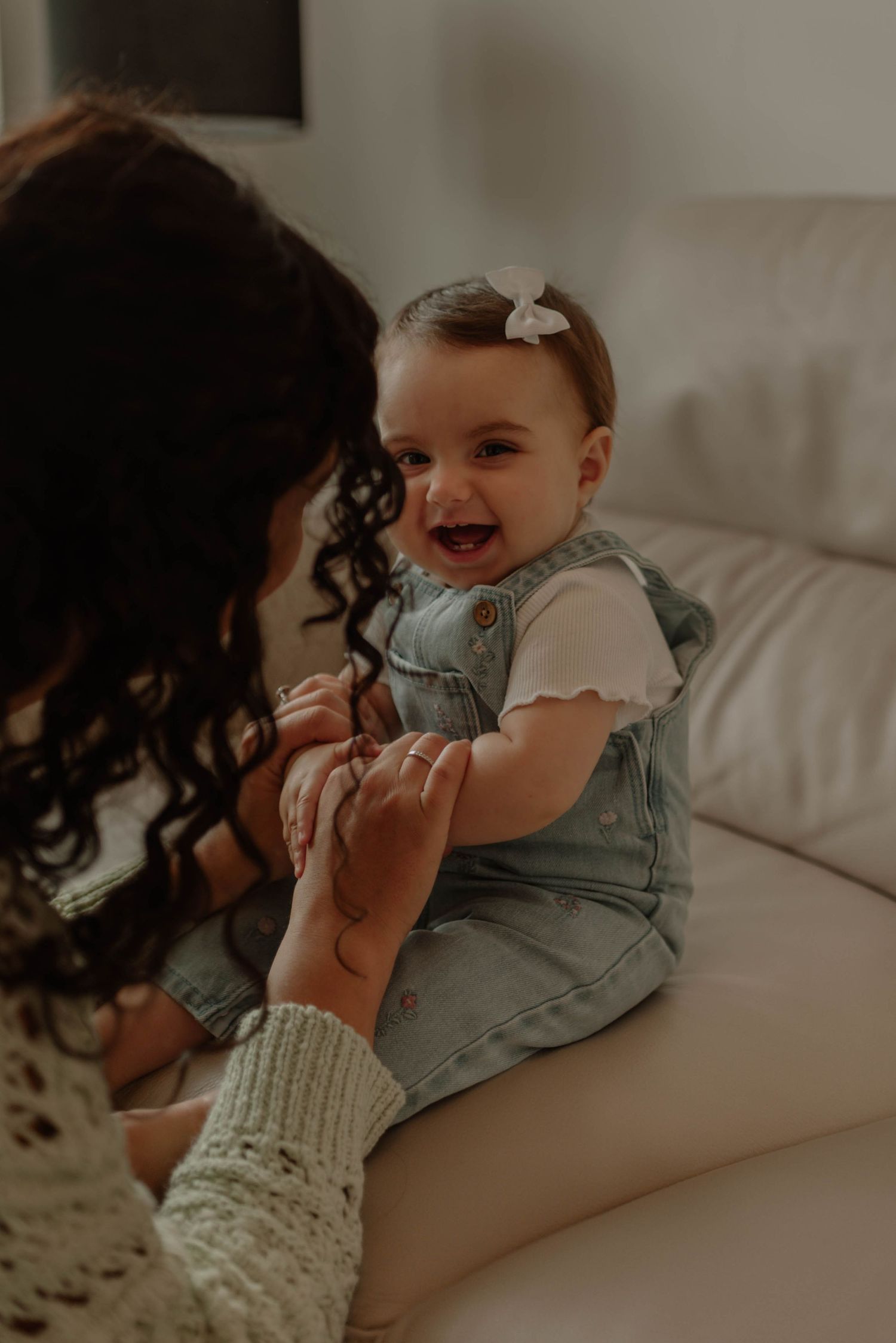 A happy baby wearing a gray dress lies on a white couch while being tickled by someone in a knit sweater.