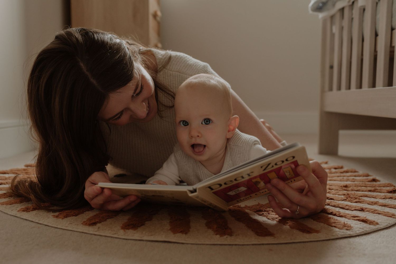 A baby lies on a round brown rug while looking up at someone leaning over them.