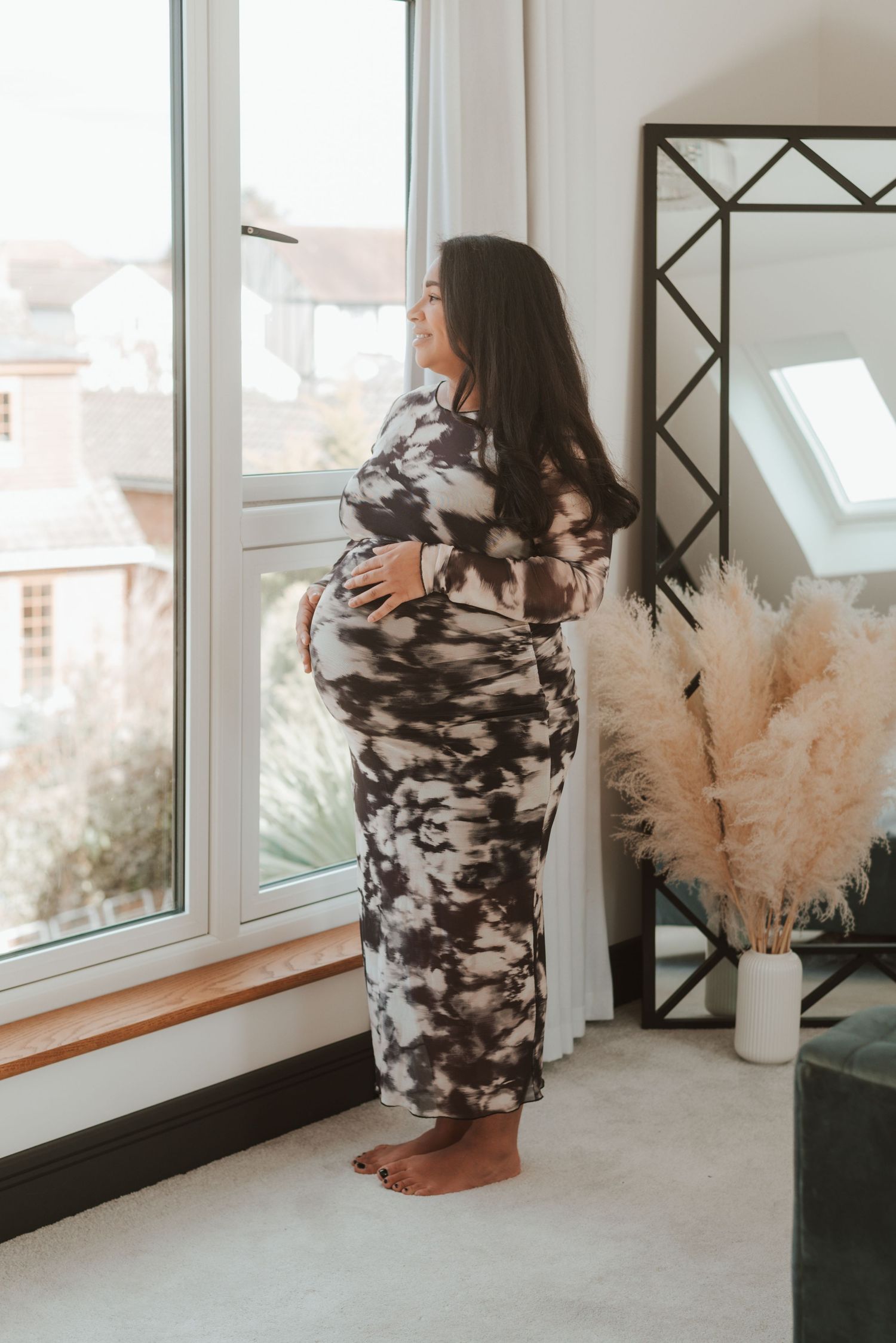 Person in a stylish black and white patterned dress standing by a window with decorative pampas grass.