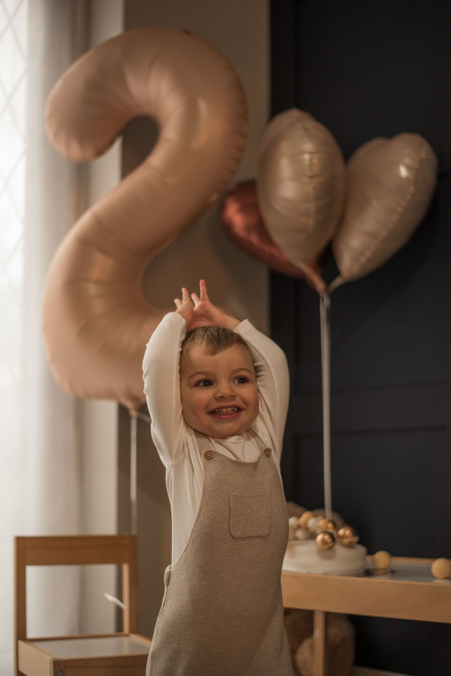 Excited toddler poses with raised arms near large number 2 balloon decoration.
