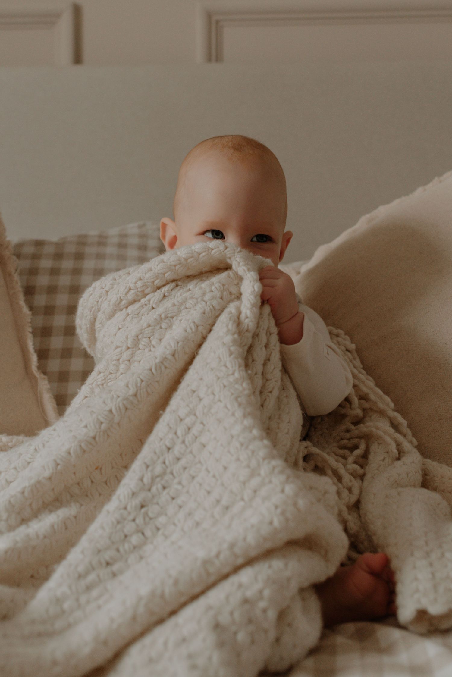 A baby peeks over a soft cream-colored blanket while lying on a couch with plaid upholstery.