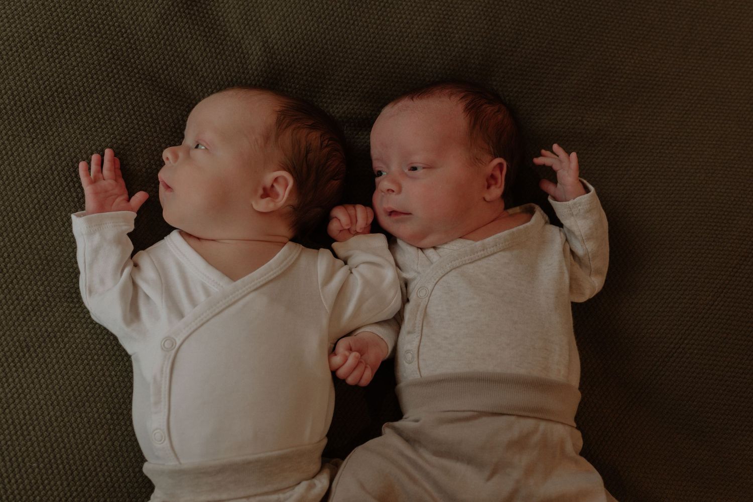 Two newborn babies in white onesies lie side by side on a dark gray surface in a series of intimate photos.